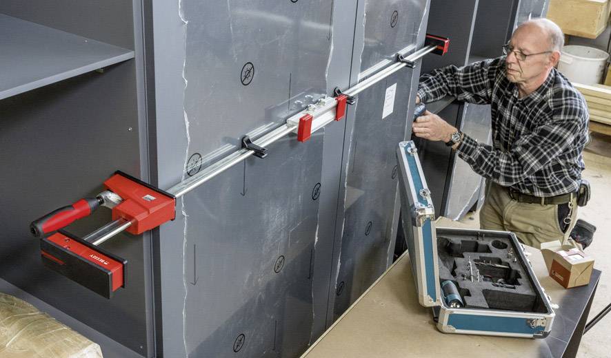 A man is securing a safe with a large red clamp. Next to him lies an open toolbox with additional accessories.