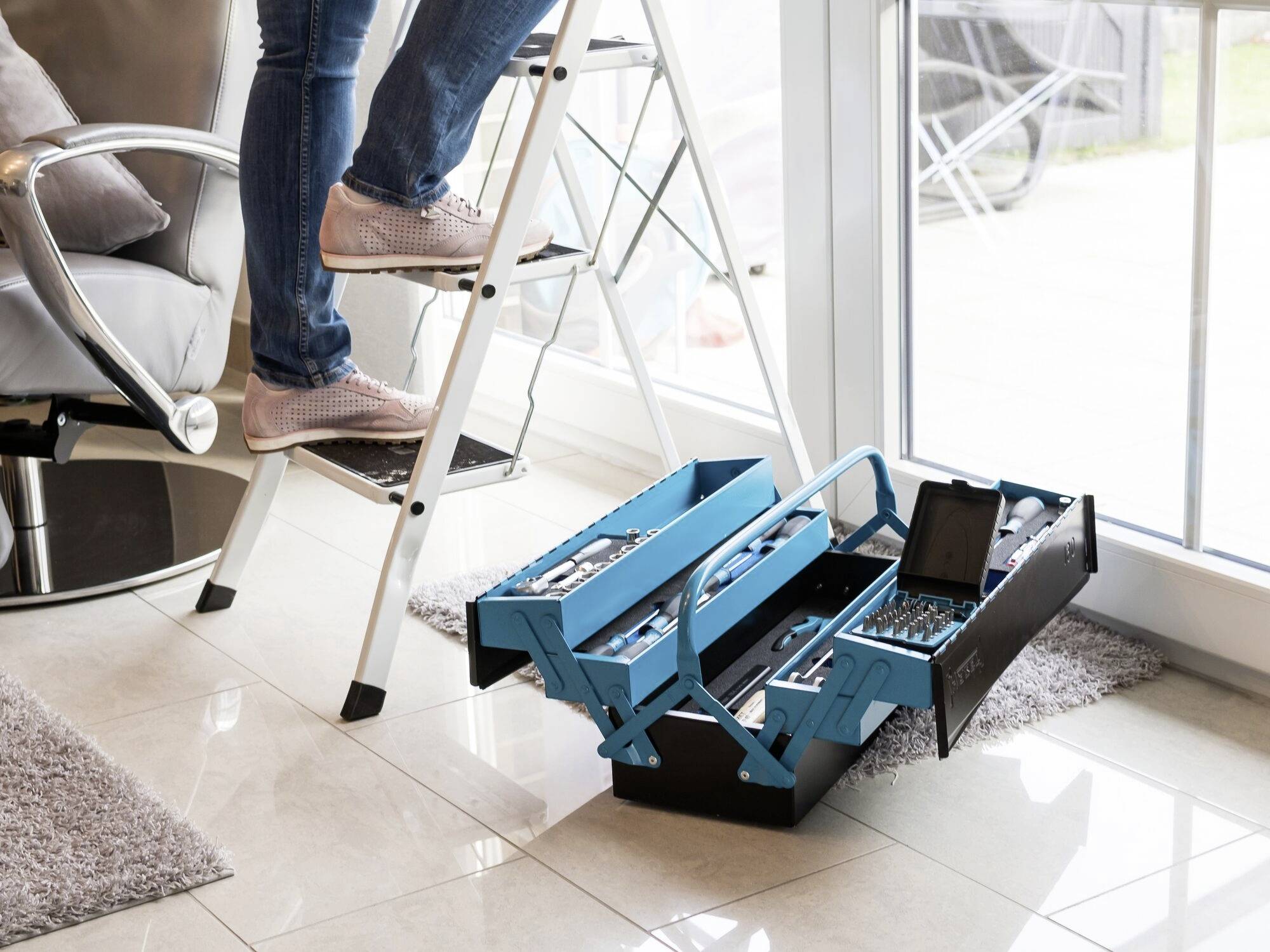 A person is climbing a ladder in a living room. An open toolbox is placed on the floor next to the ladder.
