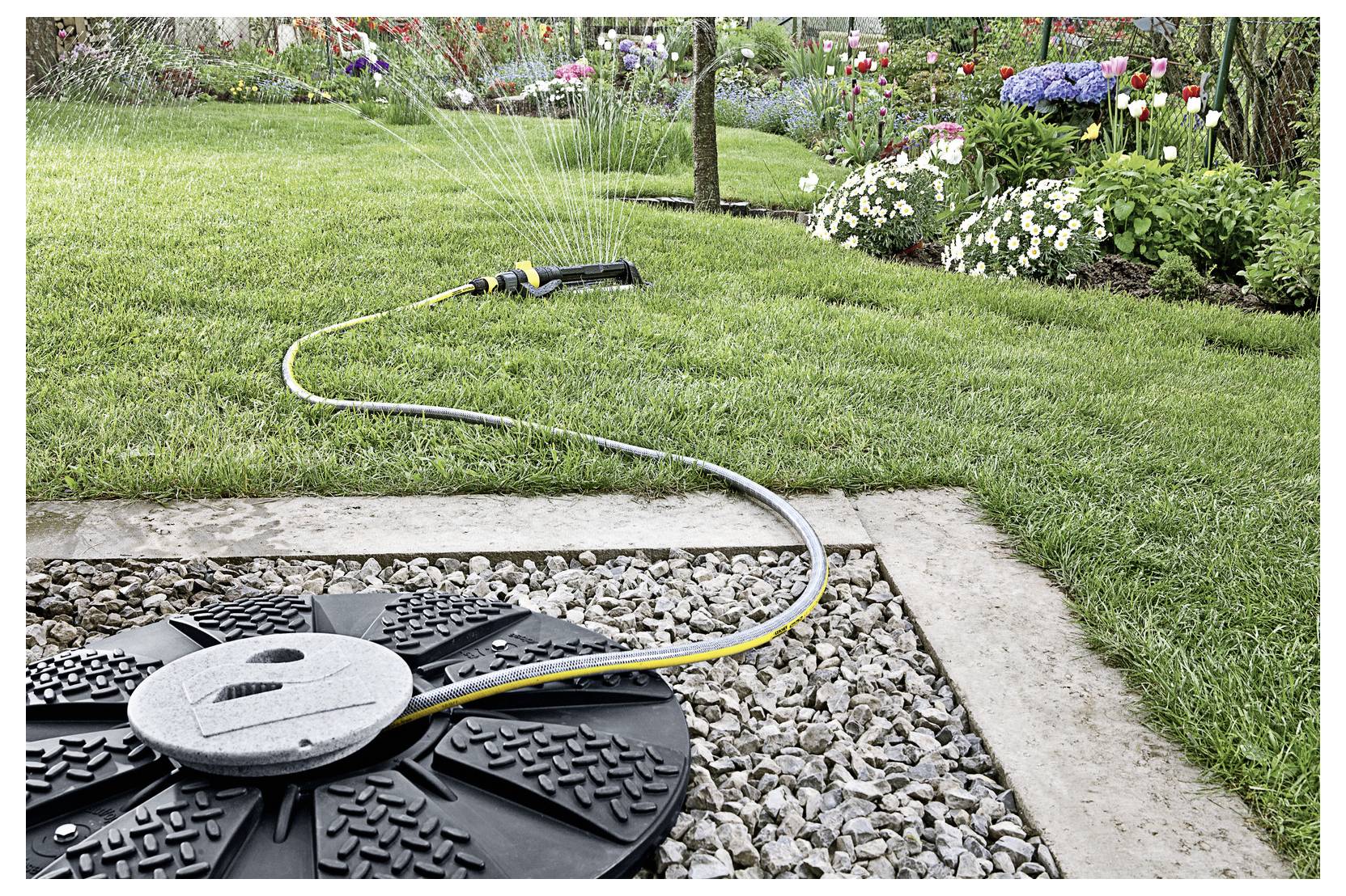 A garden sprinkler waters a lush green lawn bordered by flower beds with colorful blooms, highlighting a vibrant garden setting.