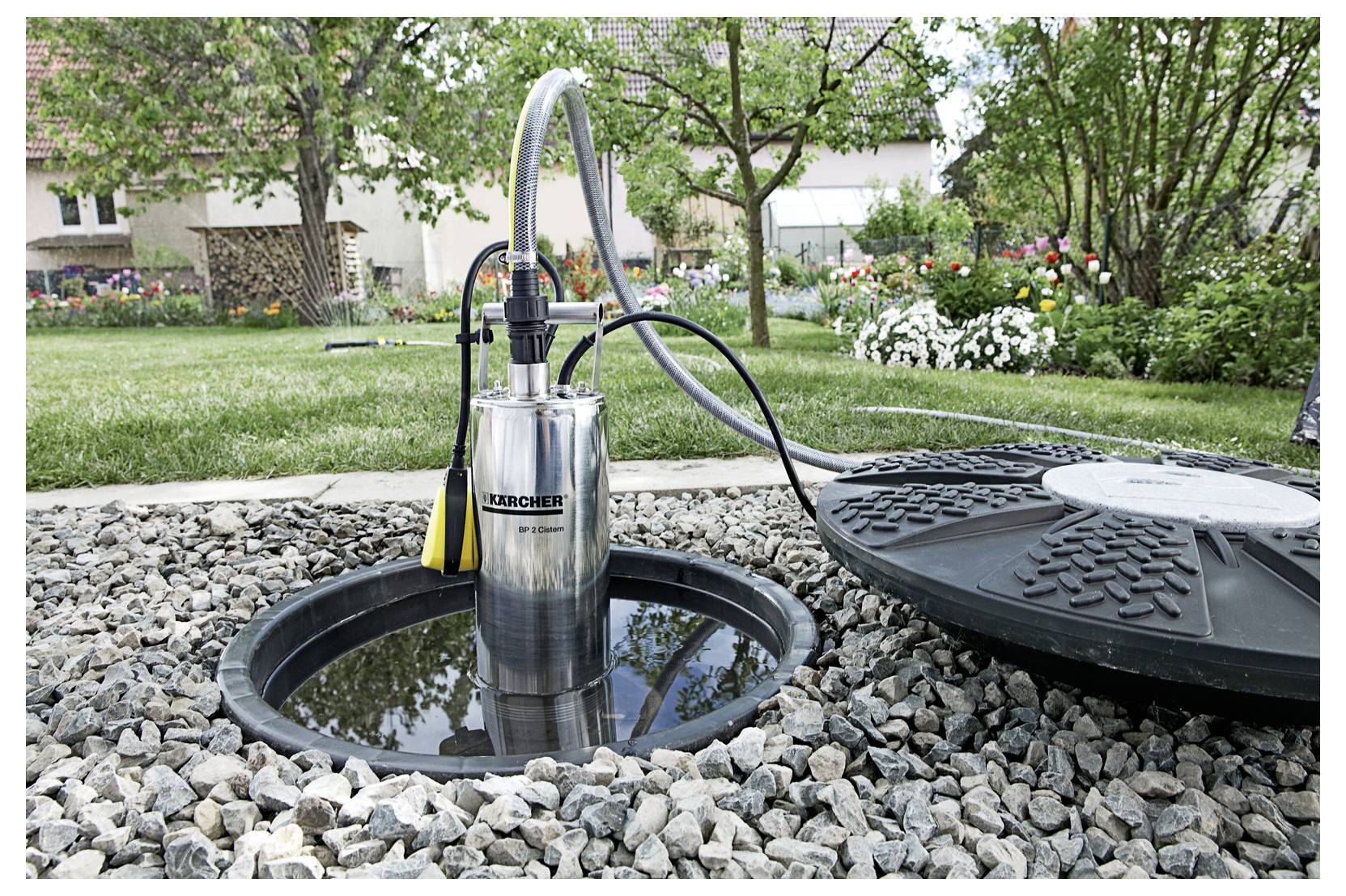 'Outdoor water pump in a pebbled garden area, surrounded by blooming flowers and trees, connected to a hose, ready for use.'