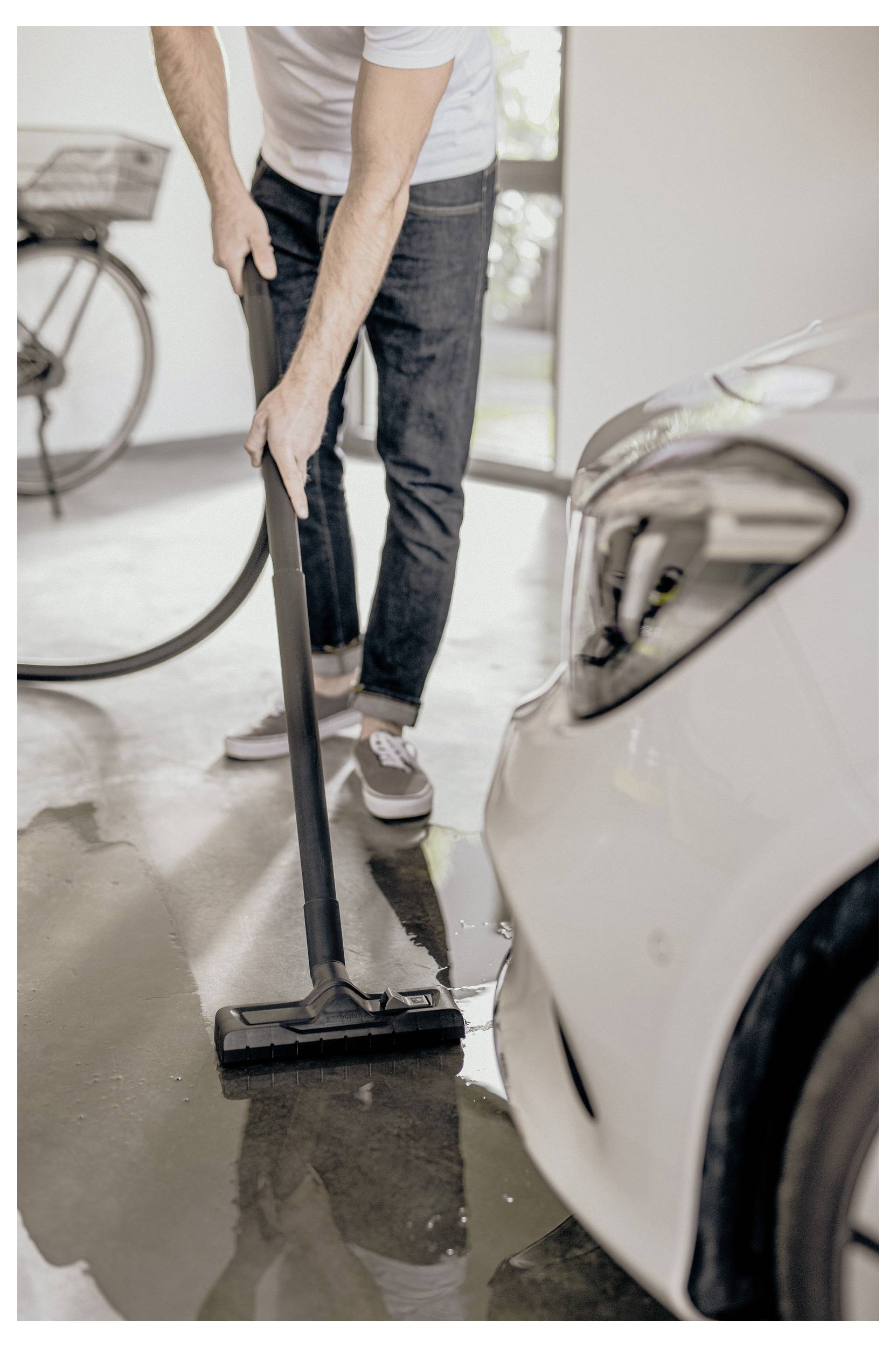 A person uses a vacuum cleaner to clean a wet garage floor next to a car, indicating tidying or maintenance activity.