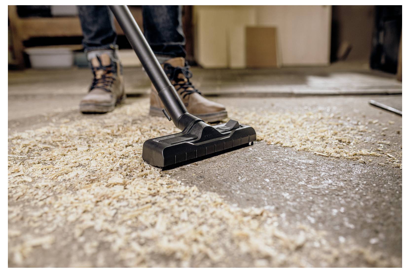 'A person uses a vacuum cleaner to remove wood shavings from a workshop floor, wearing work boots and jeans.'