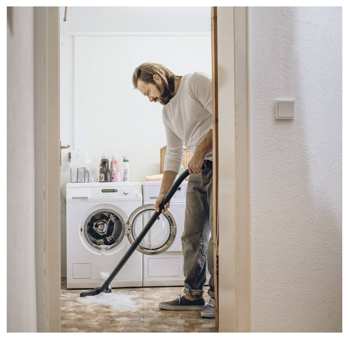 A person vacuums the floor in a laundry room with a washing machine and cleaning supplies visible in the background.