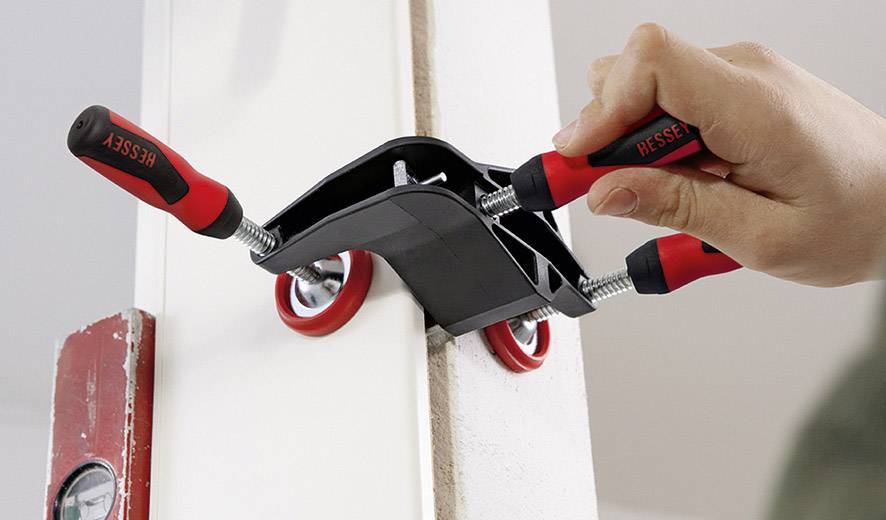 A hand is tightening two G-clamps that are securing a white board to a red beam.