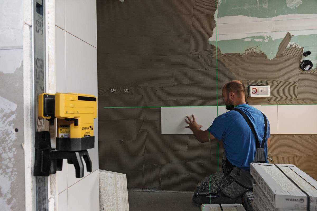 A man is installing wall tiles in a room using a laser level attached to the wall to ensure straight lines.