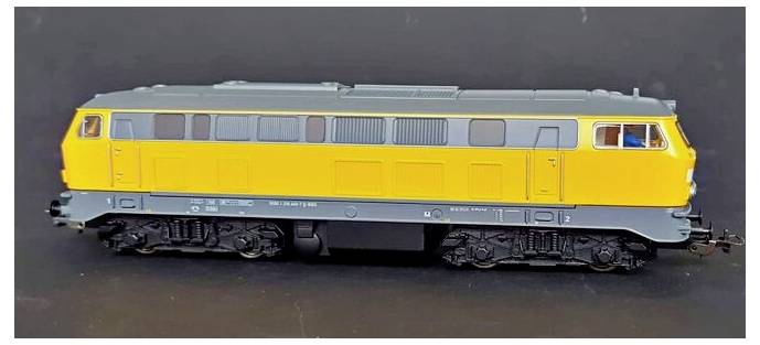 A model train locomotive painted in yellow and gray against a dark background, showing detailed features of the train's design.