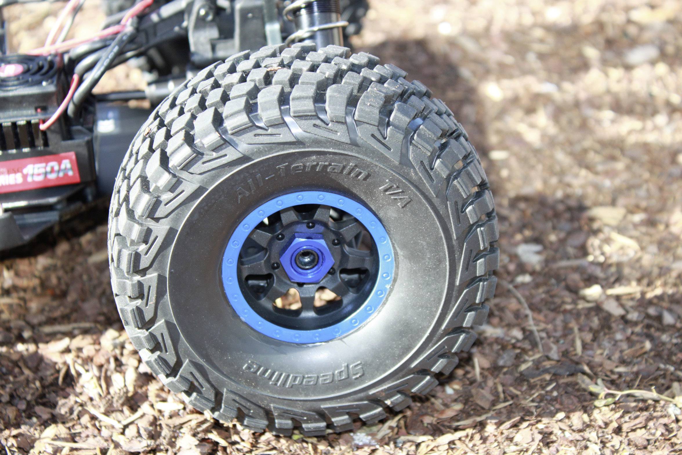 A large, robust tyre wheel of a remote-controlled car is in focus; the ground is covered with wood chippings.