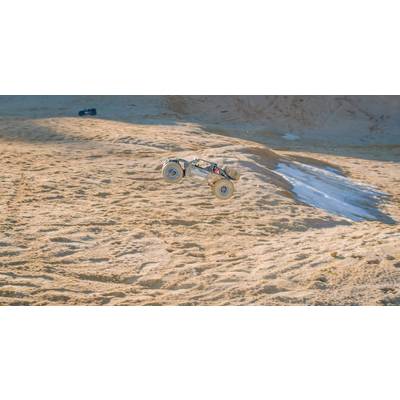 A remote-controlled car jumps through the air over a sandy dune landscape. In the background, hazy sandy ground can be seen.