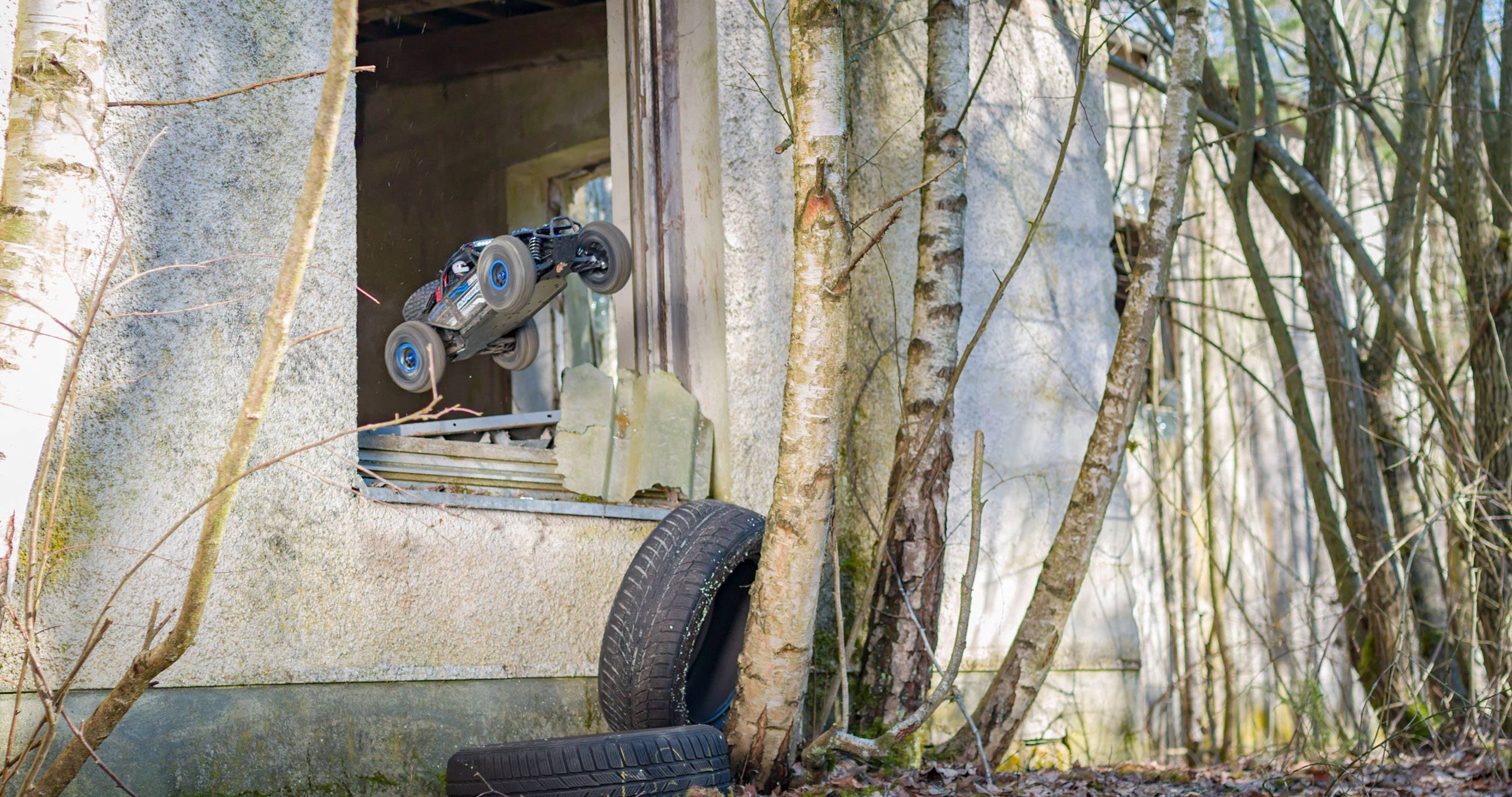 A remote-controlled car jumps through the window of an abandoned building in the woods, surrounded by trees and tyres.