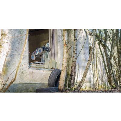 A remote-controlled car jumps through the window of an abandoned building in the woods, surrounded by trees and tyres.