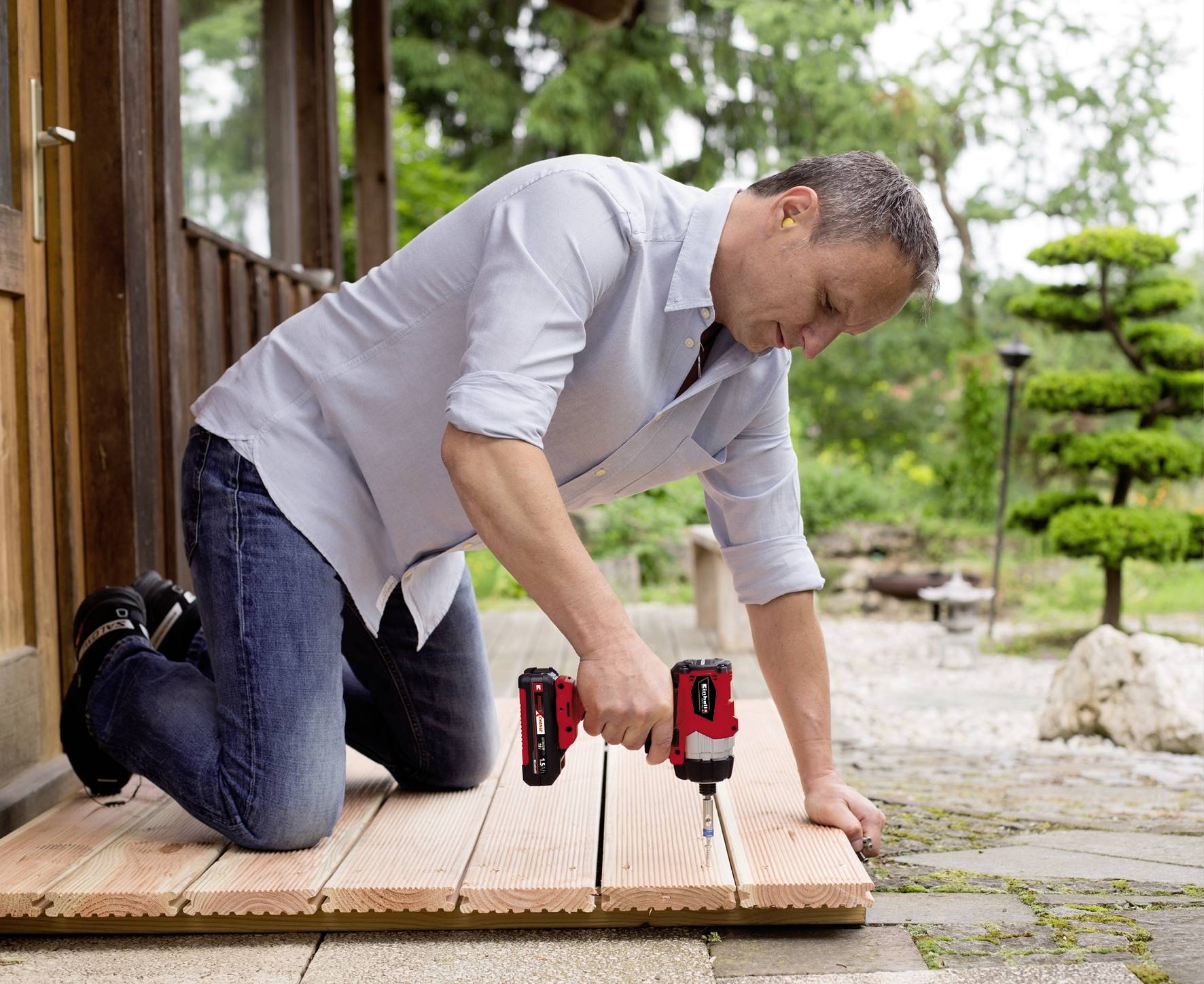 A man is kneeling outside on a terrace, securing wooden decking with a cordless drill. Trees can be seen in the background.