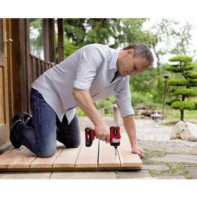 A man is kneeling outside on a terrace, securing wooden decking with a cordless drill. Trees can be seen in the background.