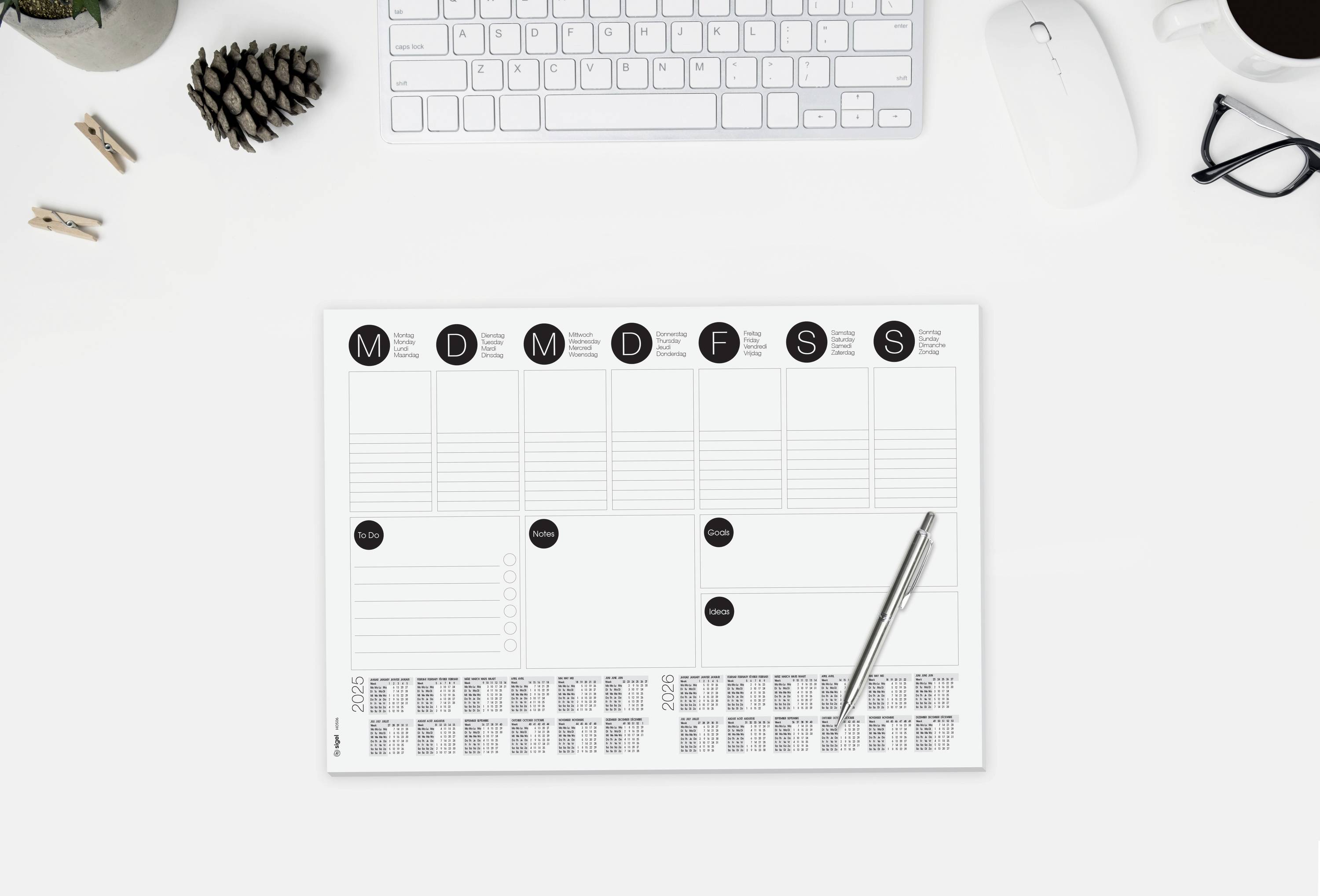 A desk with a weekly planner, pen, keyboard, mouse, glasses, cup of coffee and a small pine cone.