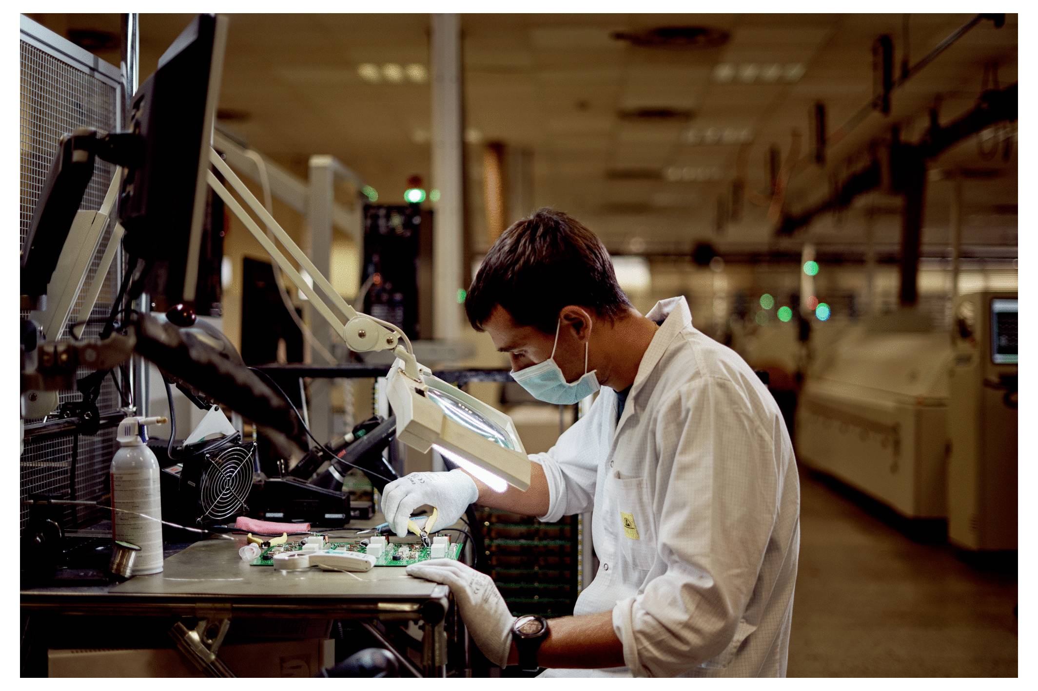 A technician in a lab coat and mask works on electronic components at a workstation in a factory setting, surrounded by equipment.