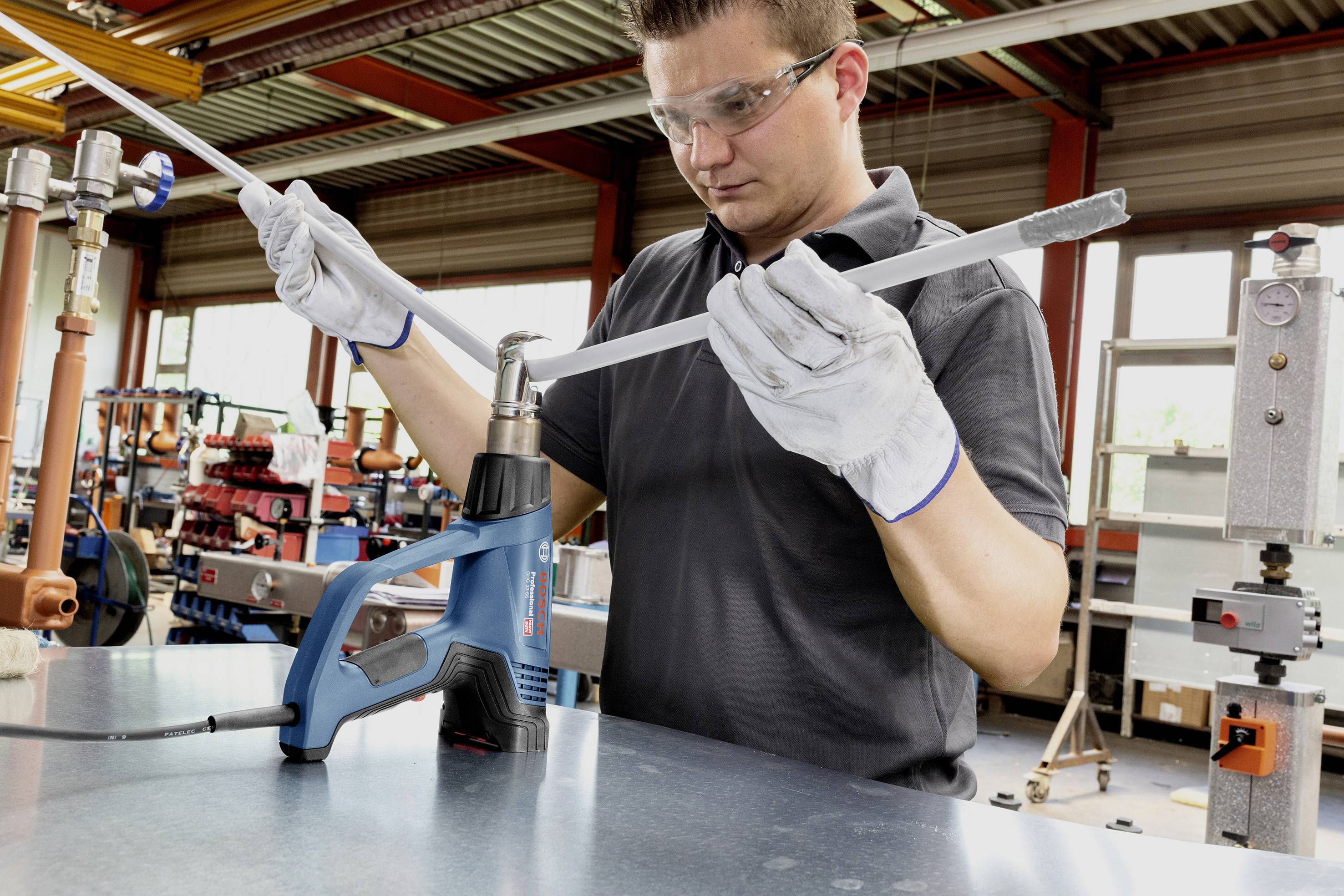 A man in work attire is using a hot air blower and gloves to work on a pipe in a workshop.
