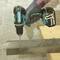 A person is drilling a hole in a metal piece with a cordless drill on a wooden table. The background is a concrete wall.