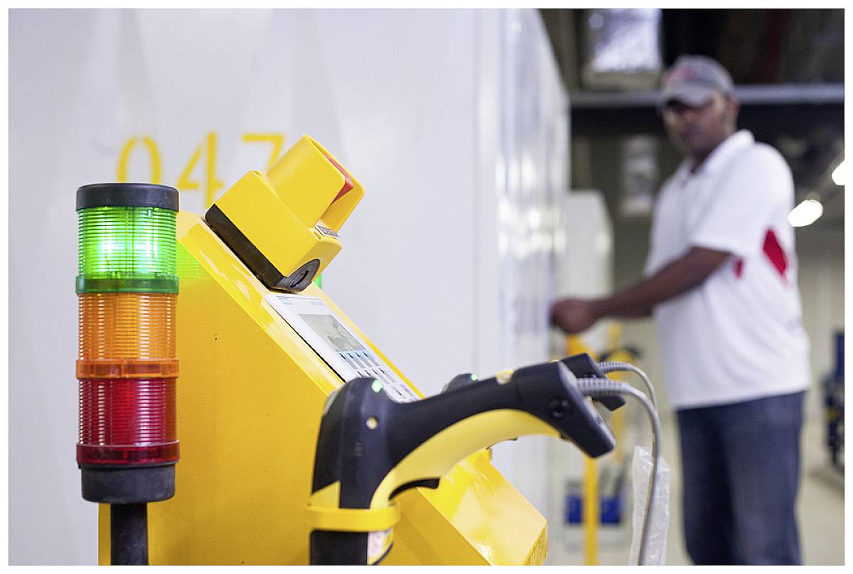 A person in the background is operating a device in a factory. In the foreground, a machine with green, yellow, and red lights displays operational status.