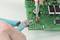Close-up of hands working on an electronic circuit board; pliers and tweezers holding a red LED in position.