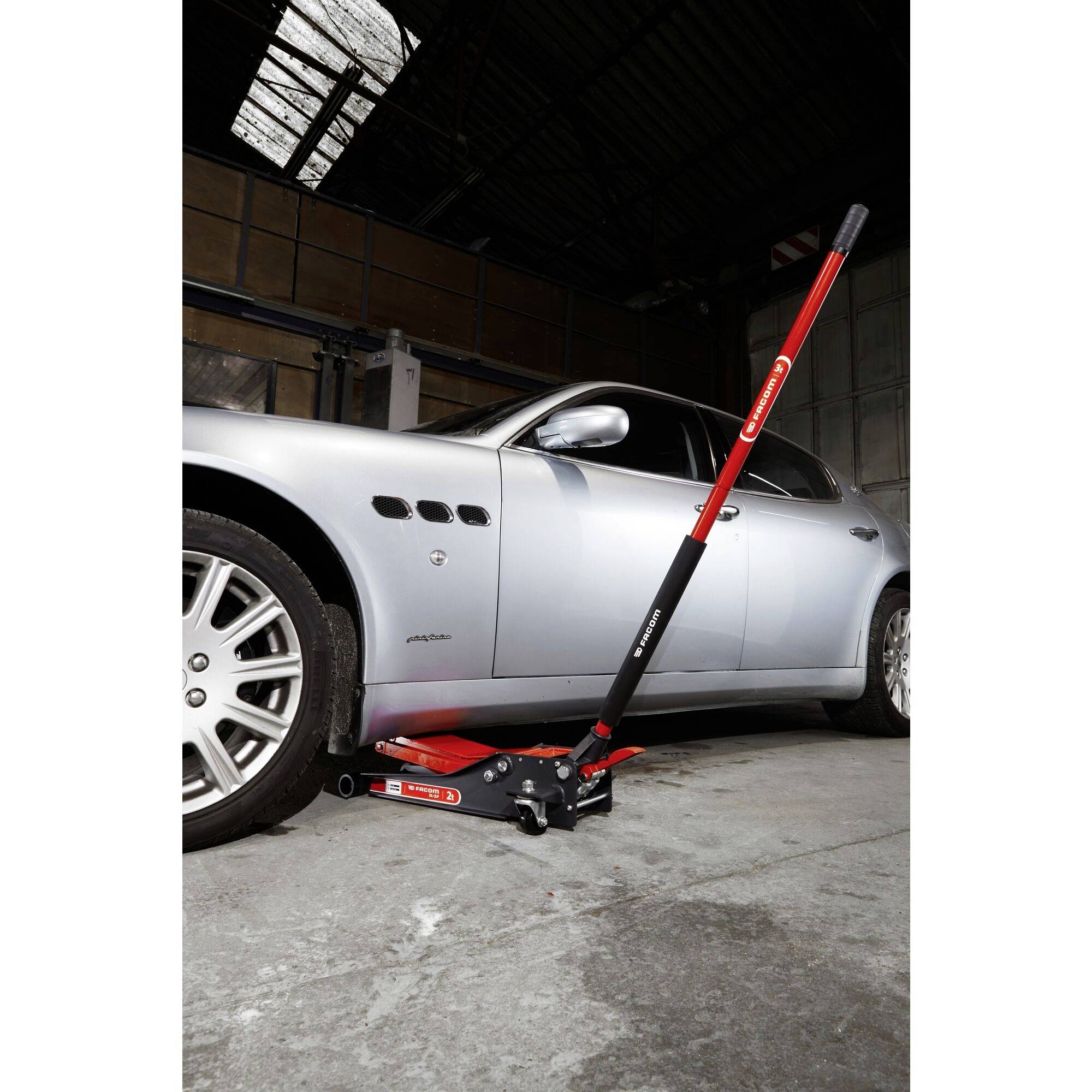A silver car is being raised by a red car jack in a workshop. Workshop walls and a roof window can be seen in the background.