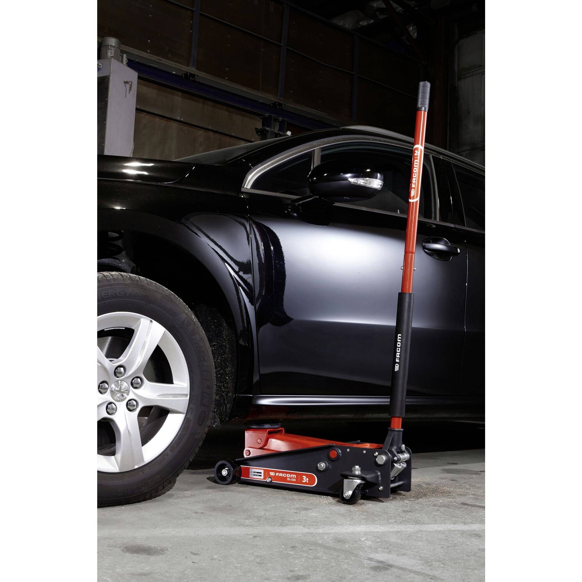 A black car is being raised by a red hydraulic jack in a workshop. The jack is marked with the 'Husky' label.