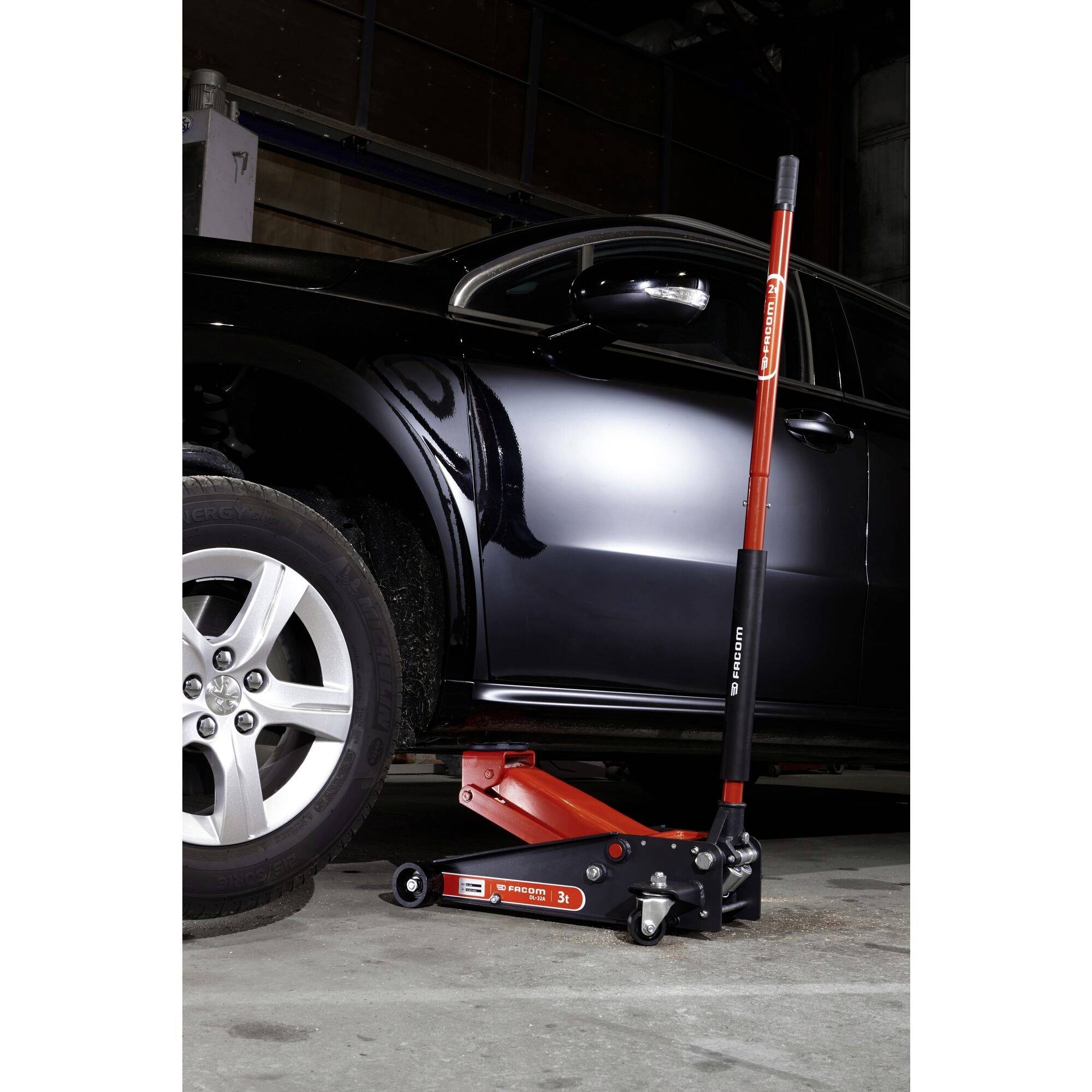 A car is being lifted with a hydraulic jack. The jack is red and is standing on a concrete floor. The car has a black paint finish.