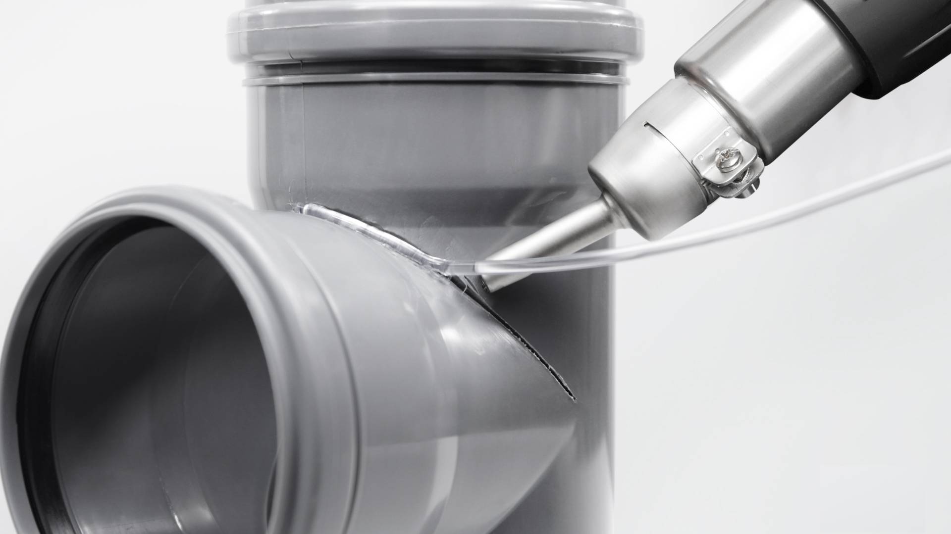 A metal tool is welding plastic pipes together. One pipe is angled at a right angle to the camera, while the tool processes the seam.