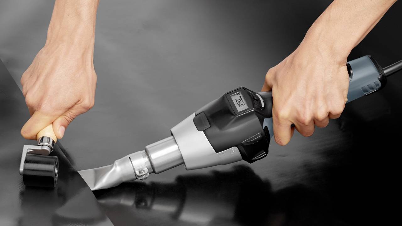 Two hands are working with a hot air blower and a roller tool on black material, presumably for sealing or welding work.