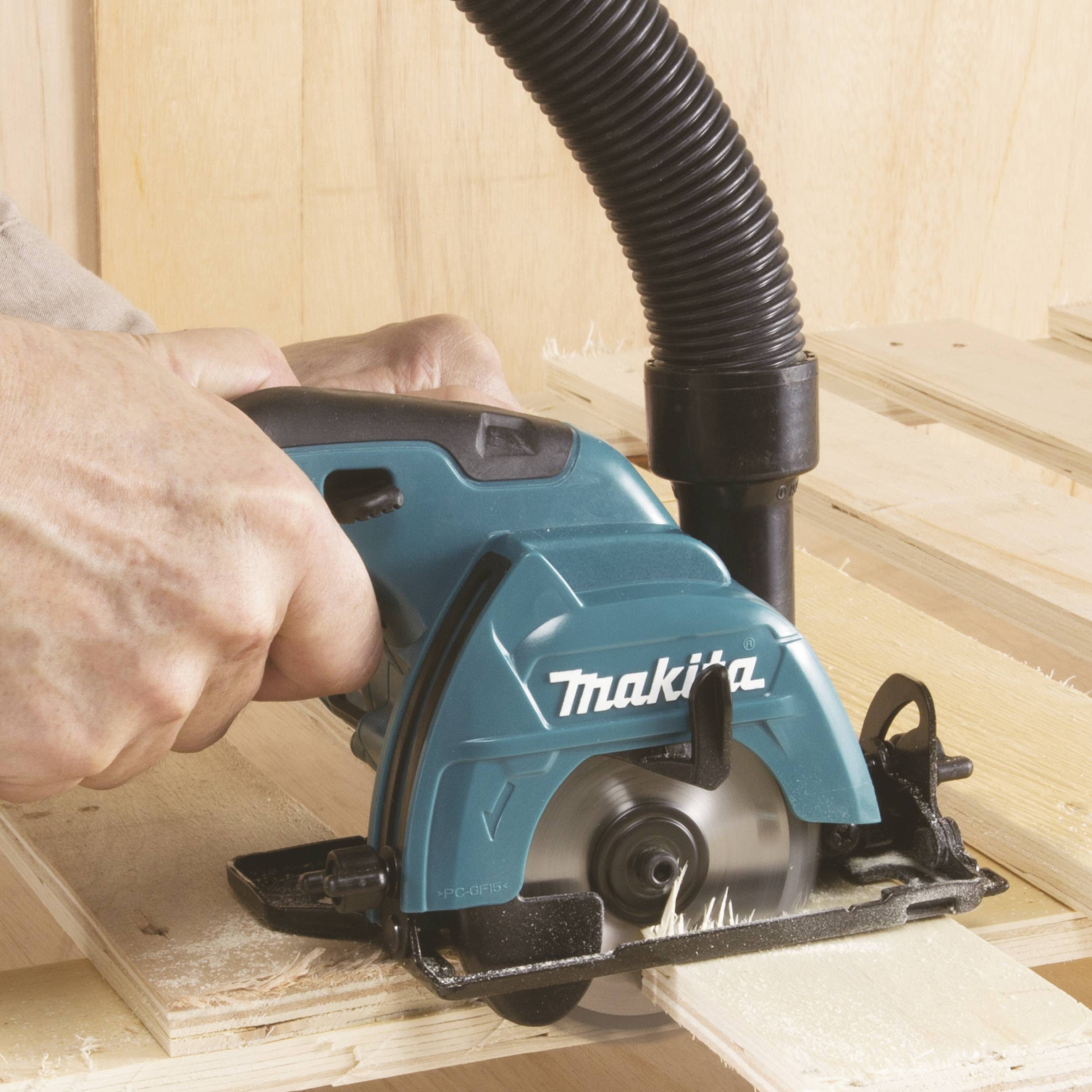 A hand is holding an electric saw cutting wood. Dust is being extracted by an attached hose.