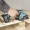 A hand guides a Makita cordless circular saw over a piece of wood. The saw is cutting through the board.