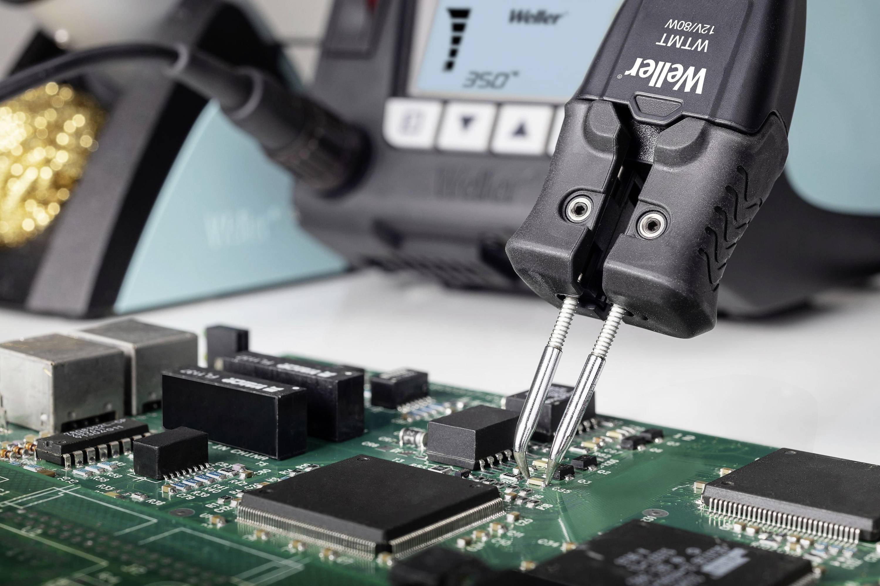 Close-up of a circuit board with a soldering iron working on electronic components. In the background, a soldering iron stand is positioned.