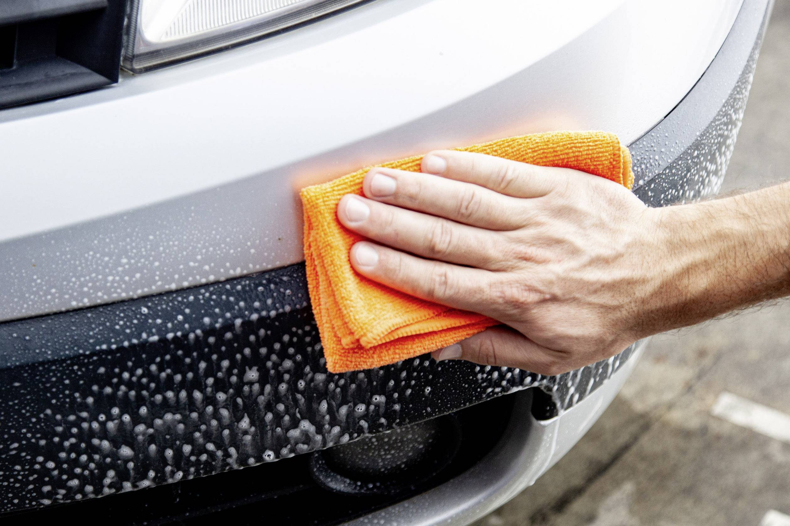 'A hand is cleaning the wet, soapy car at the bumper with an orange cloth.'