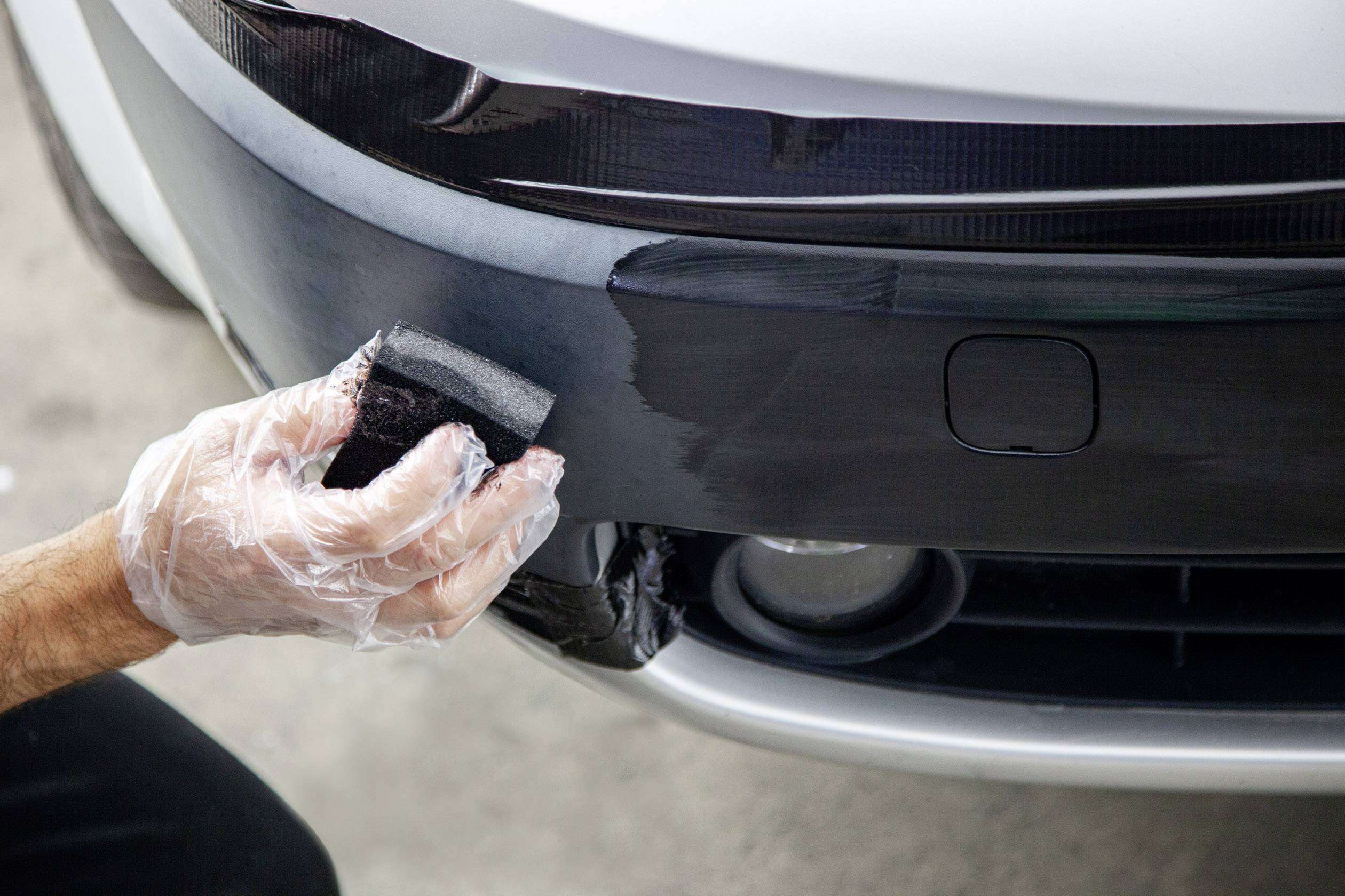 A person is polishing the front bumper of a car with a sponge. The hand is wearing a disposable glove.