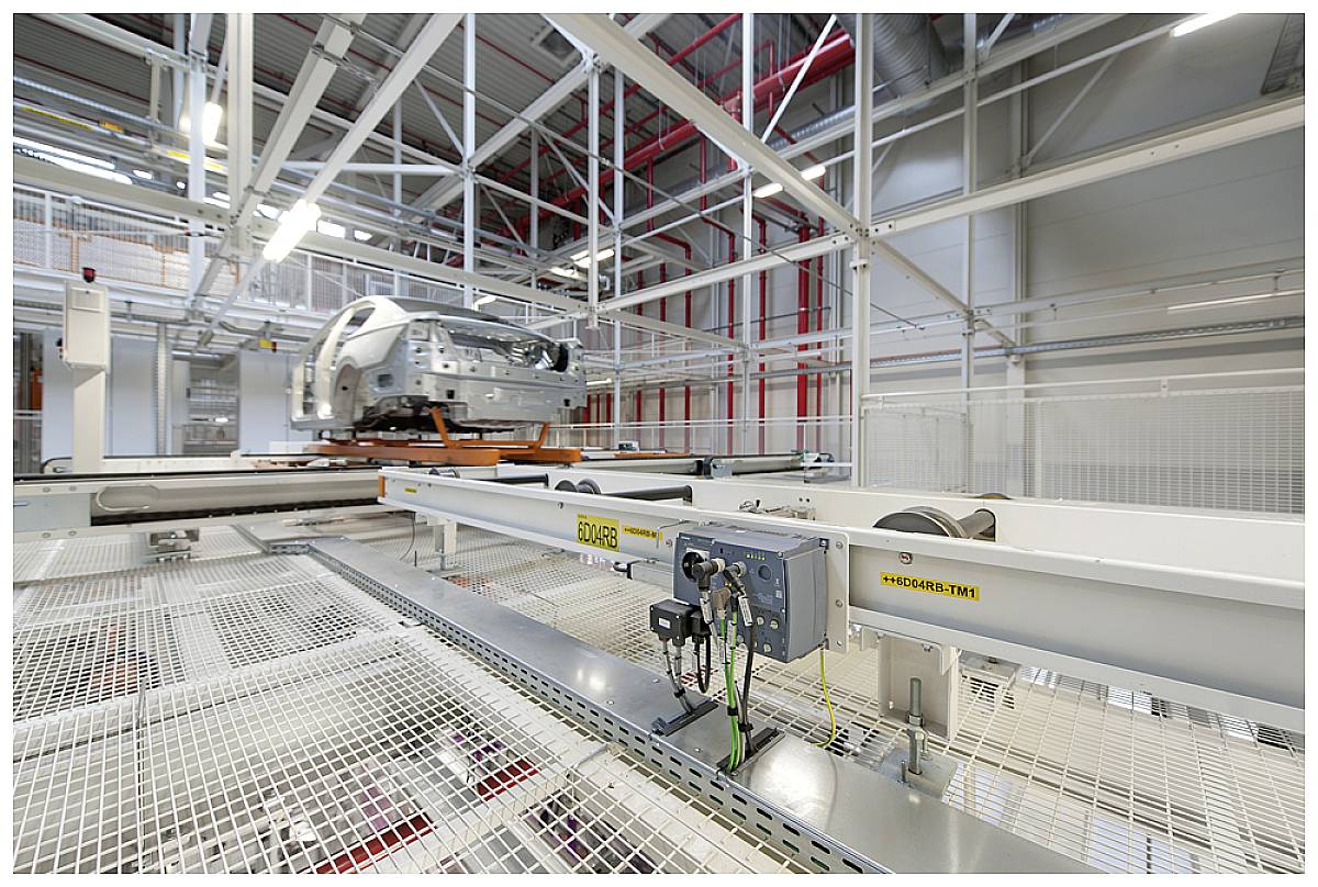 Factory hall with a car on a lifting device, surrounded by steel beams and wire mesh. Industrial environment, well-lit.