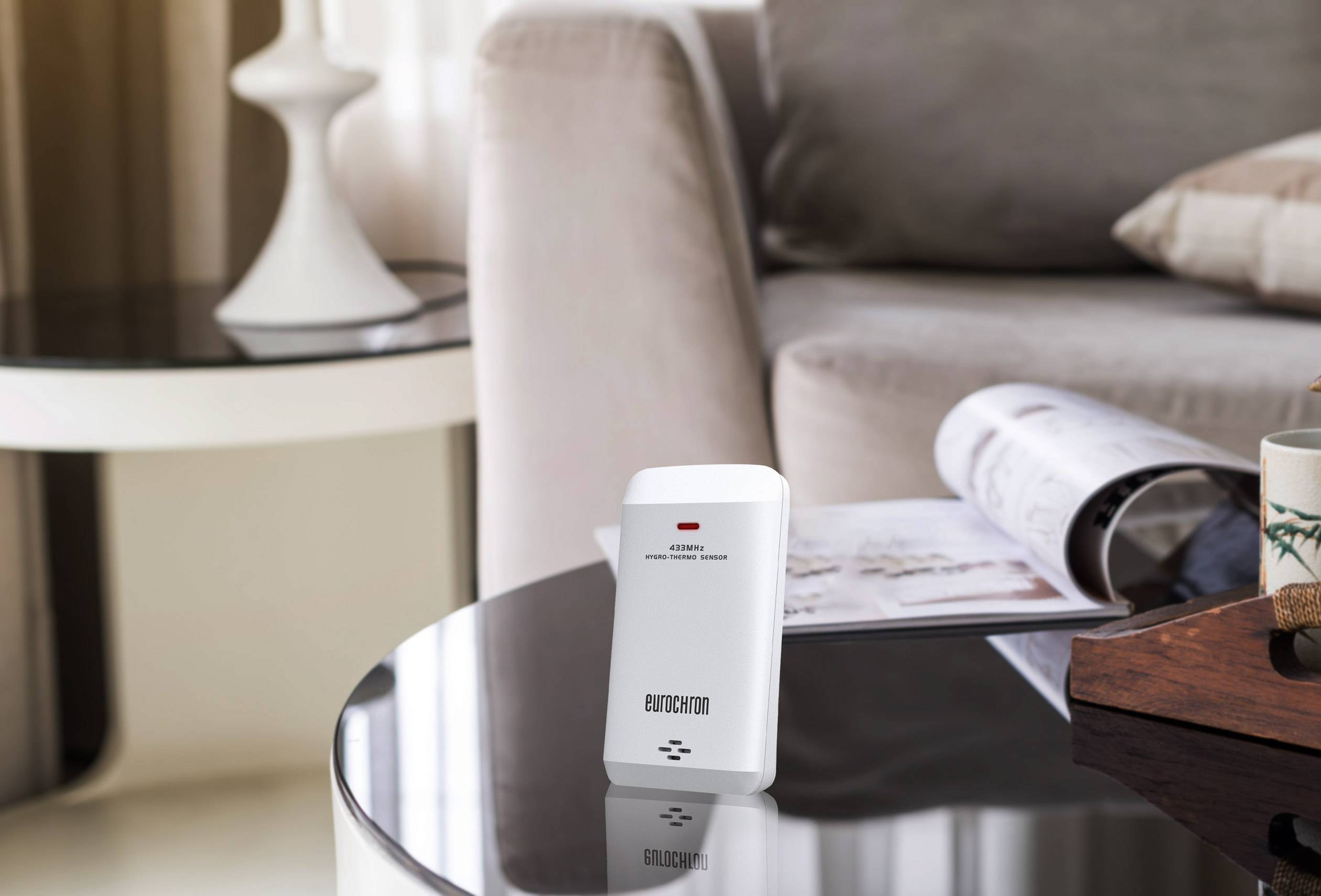 A motion sensor sits on a glass table in the living room. In the background, a sofa, a cushion, and an open magazine can be seen.