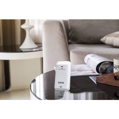 A motion sensor sits on a glass table in the living room. In the background, a sofa, a cushion, and an open magazine can be seen.