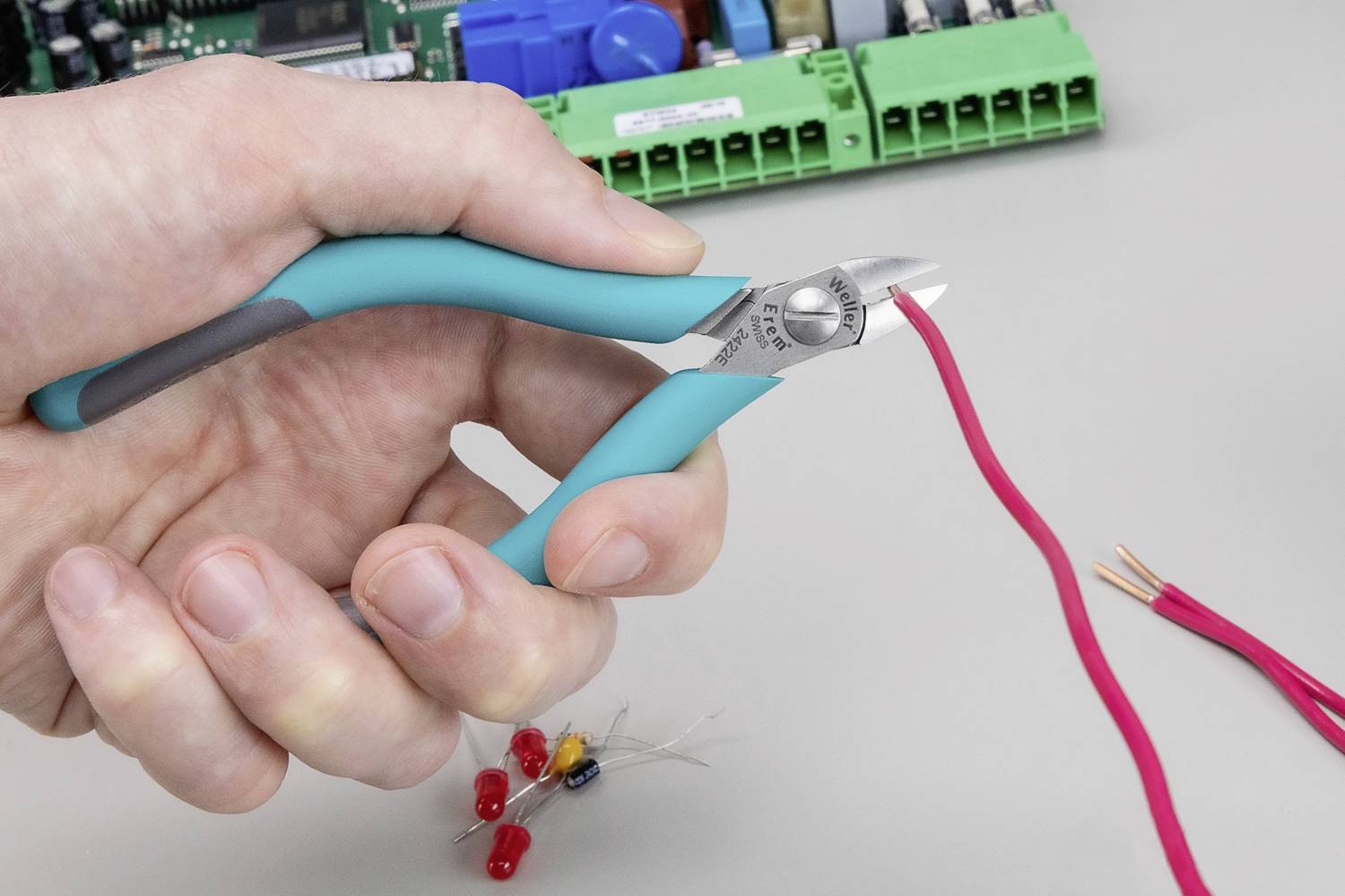 A hand is holding wire cutters and cutting a red cable. A circuit board is visible in the background.