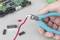 A hand holds wire cutters and cuts a red cable. Electronic components and a circuit board are lying in the background.