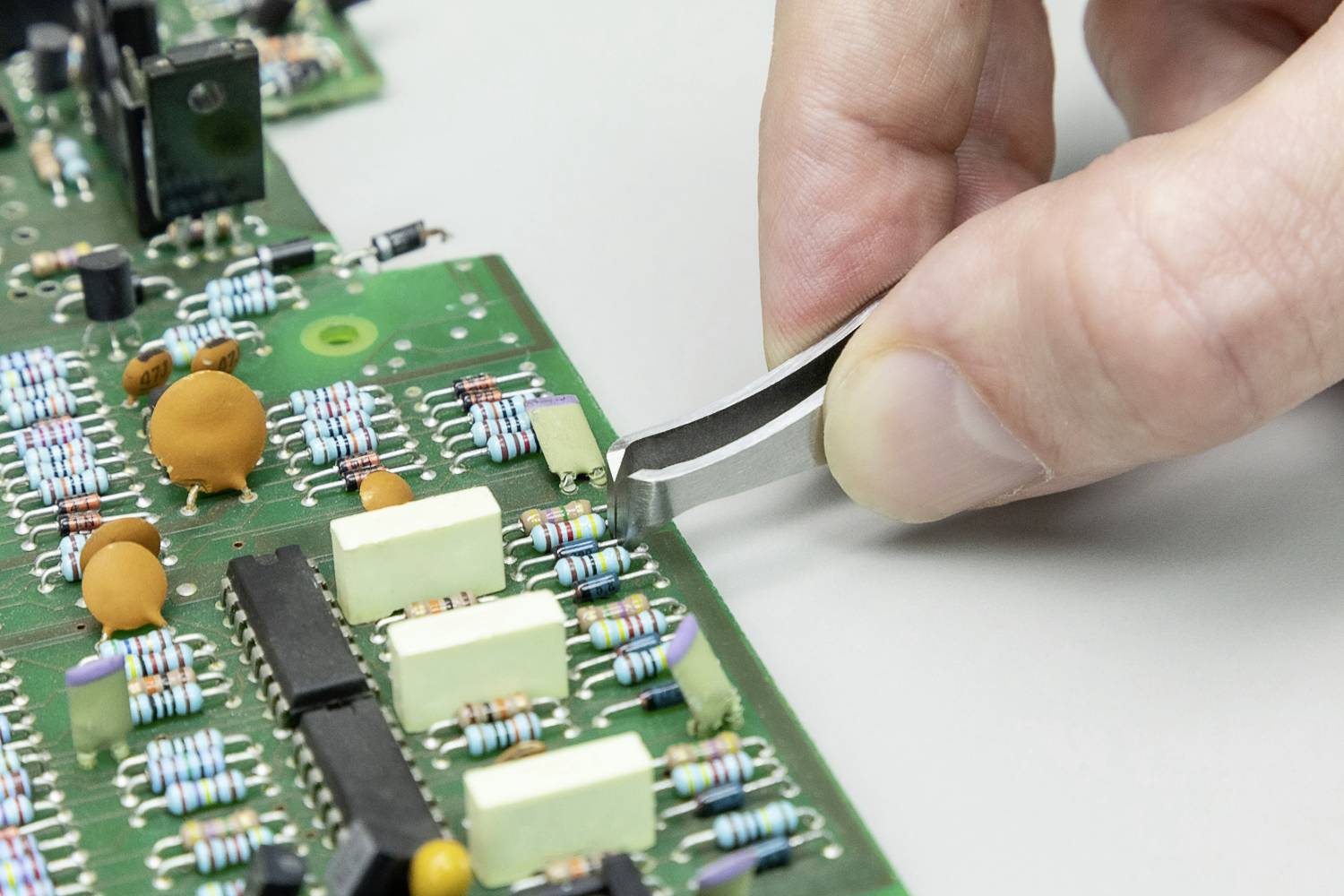 A hand uses tweezers to place a small component on a green circuit board. Various resistors and capacitors are visible.