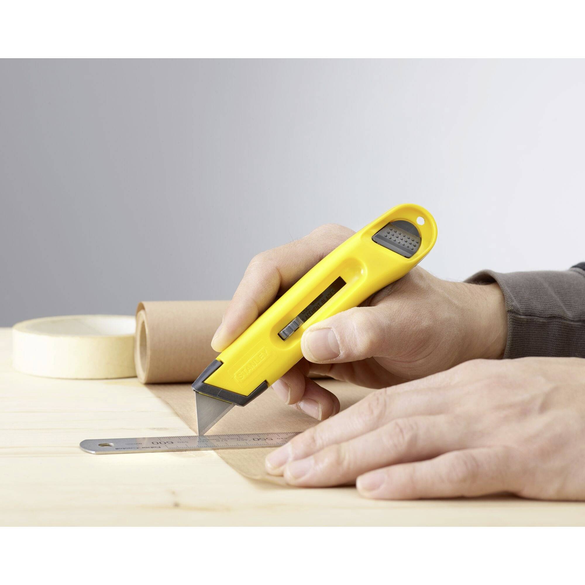 A person is cutting with a yellow utility knife along a metal ruler on a wooden table. Rolls of adhesive tape are visible in the background.