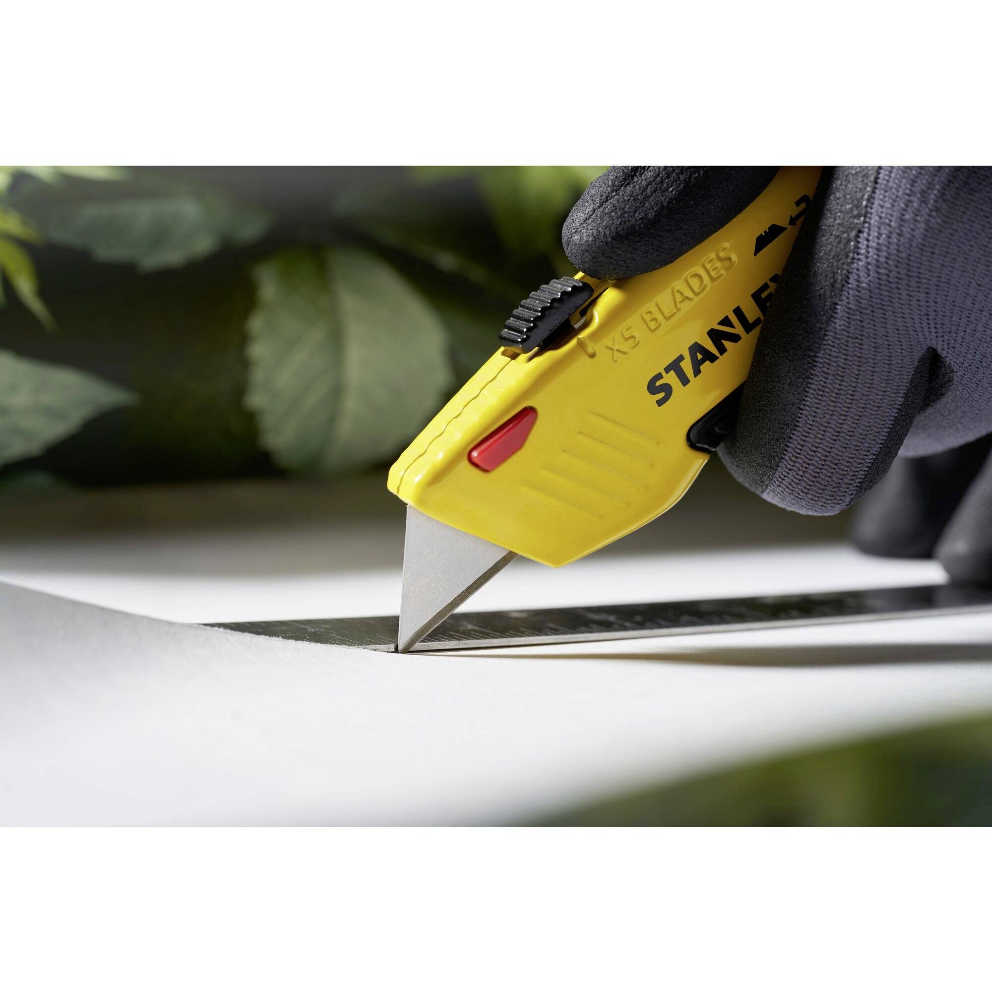 A hand wearing a black glove is using a yellow utility knife to cut a white material.