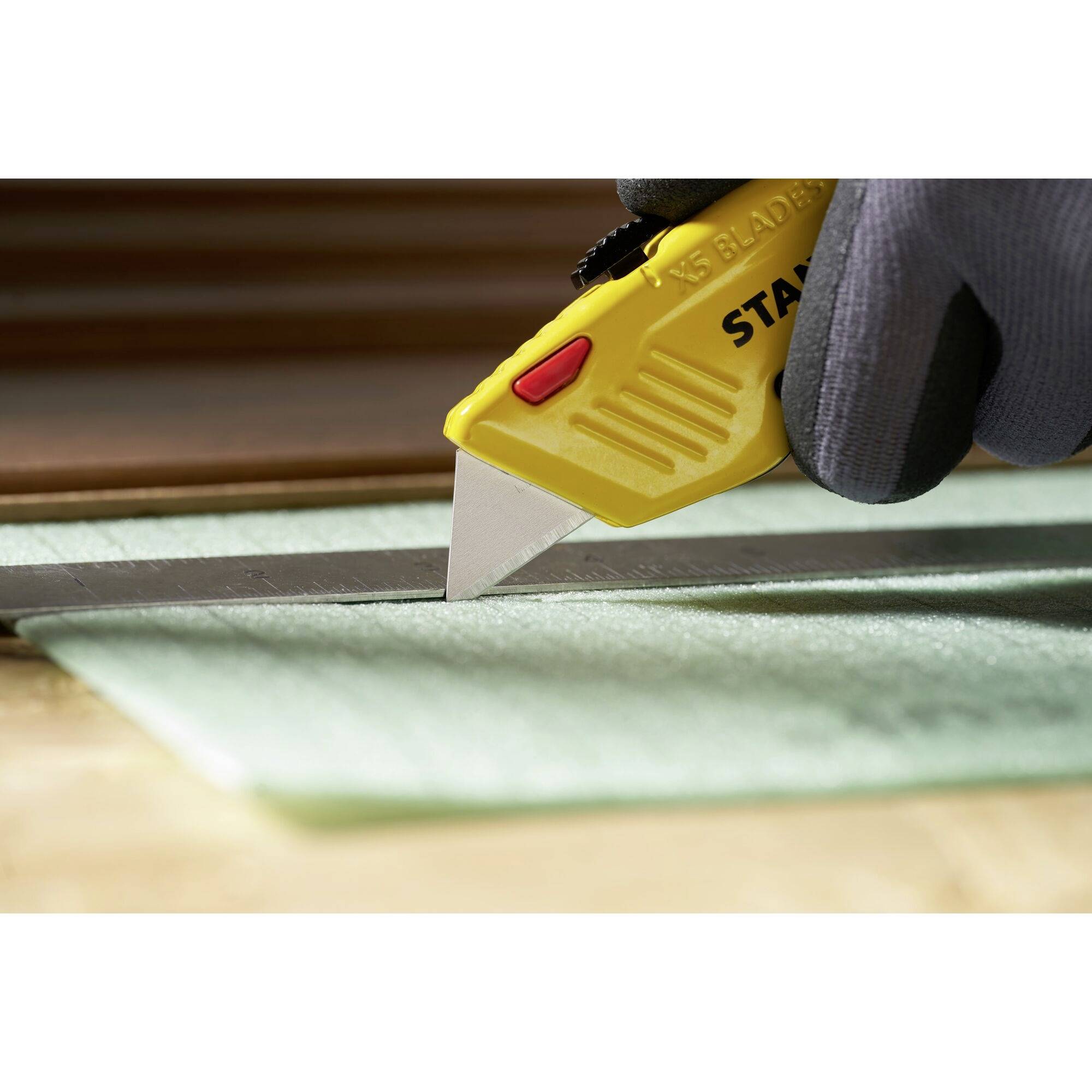 A gloved hand holds a yellow craft knife and is cutting a green material with the help of a ruler.