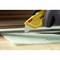 A gloved hand holds a yellow craft knife and is cutting a green material with the help of a ruler.