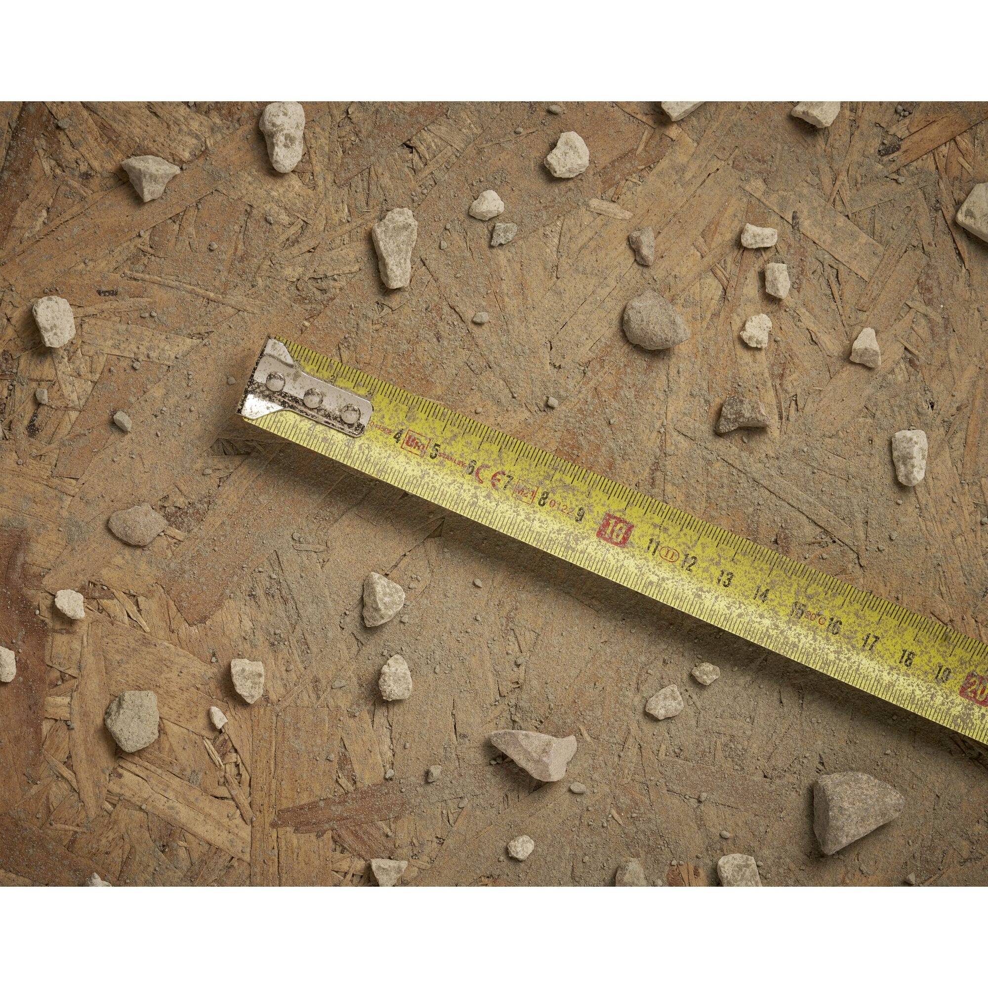 A yellow measuring tape lies diagonally on a wood-like surface with scattered small stones, creating the impression of a construction site.