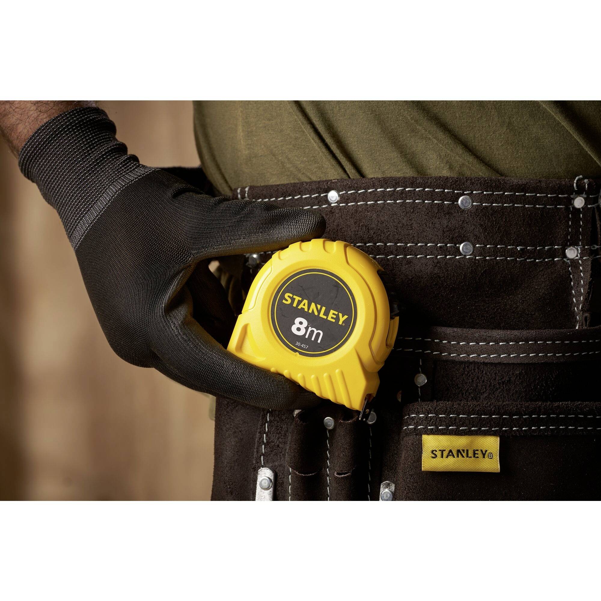 'A man in work gloves is putting a yellow 8m Stanley tape measure into a work bag.'