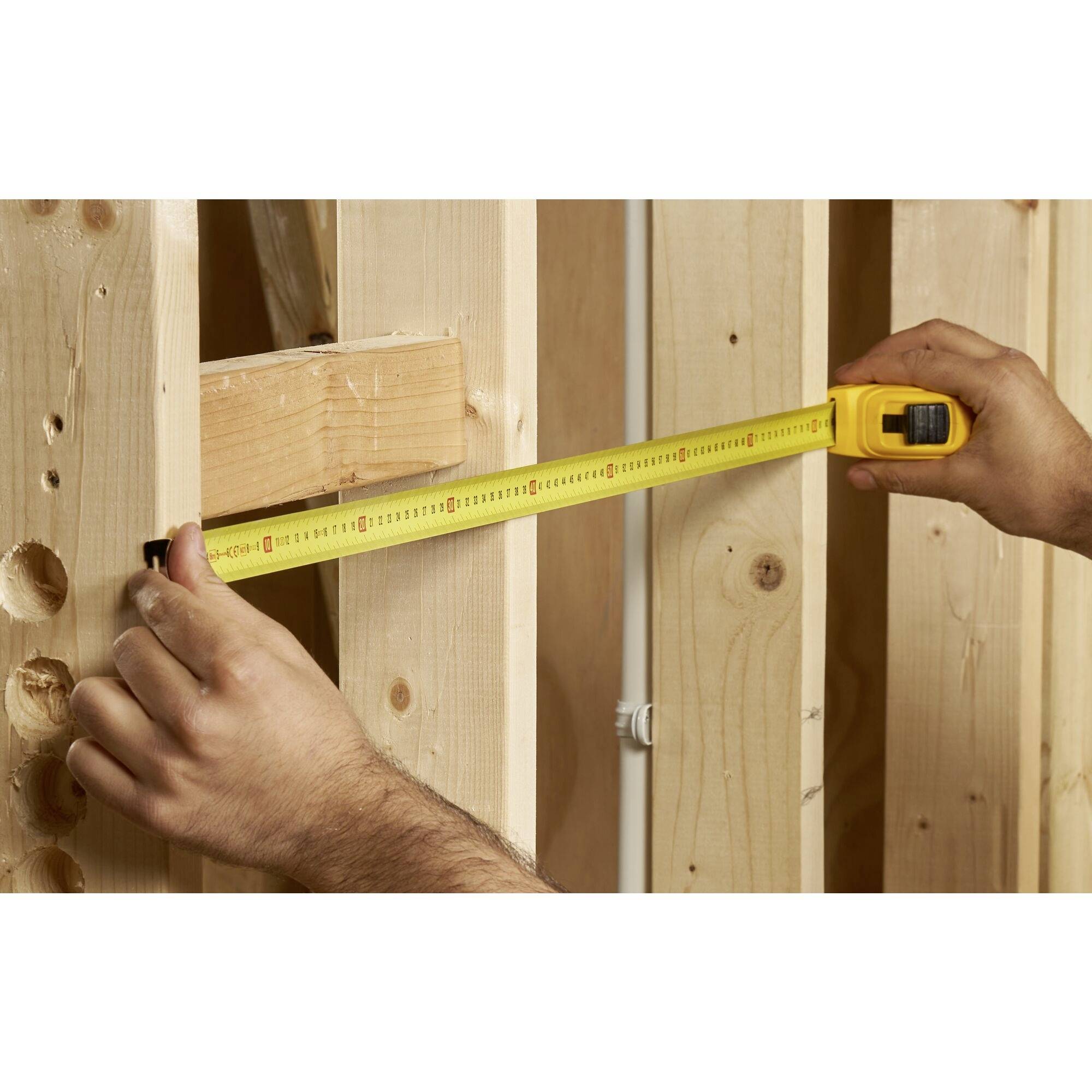 Someone is measuring the distance between two wooden beams in an unfinished building using a yellow measuring tape.