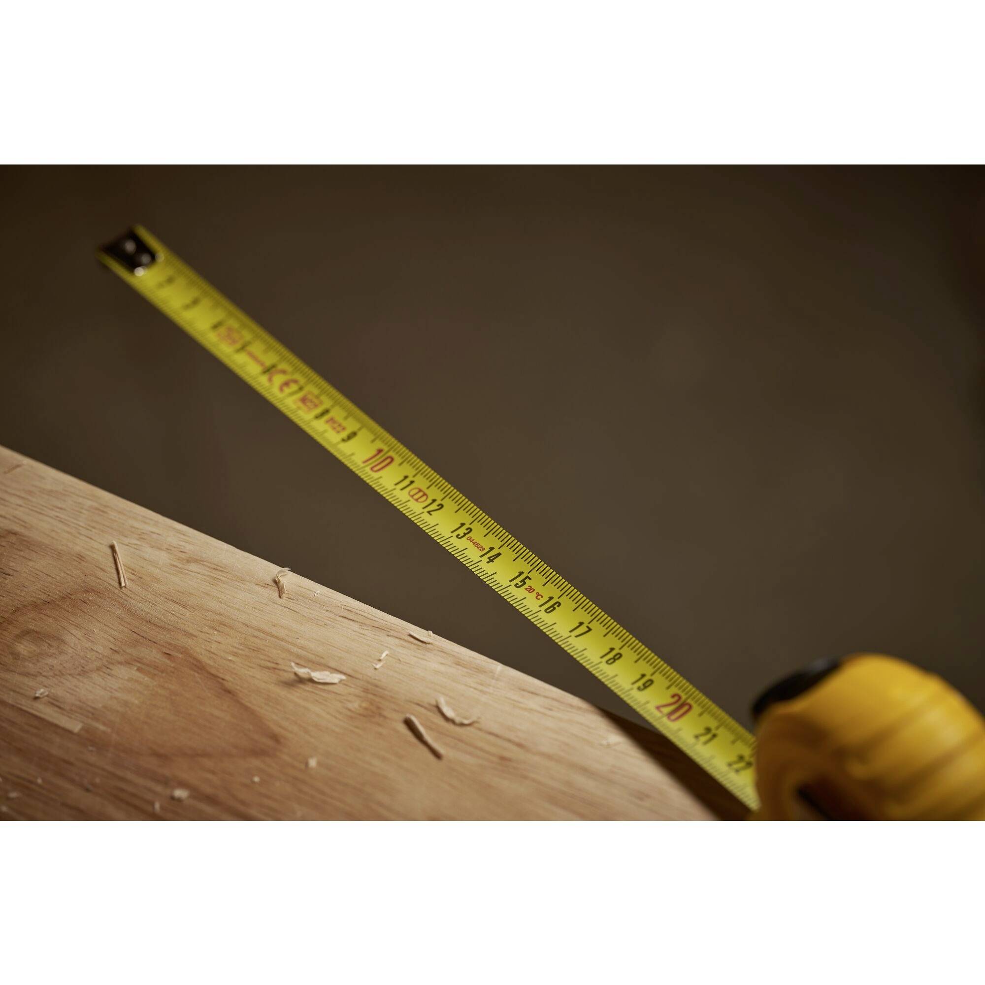 A yellow tape measure stretches across a wooden board with visible nails and wood shavings in the foreground.