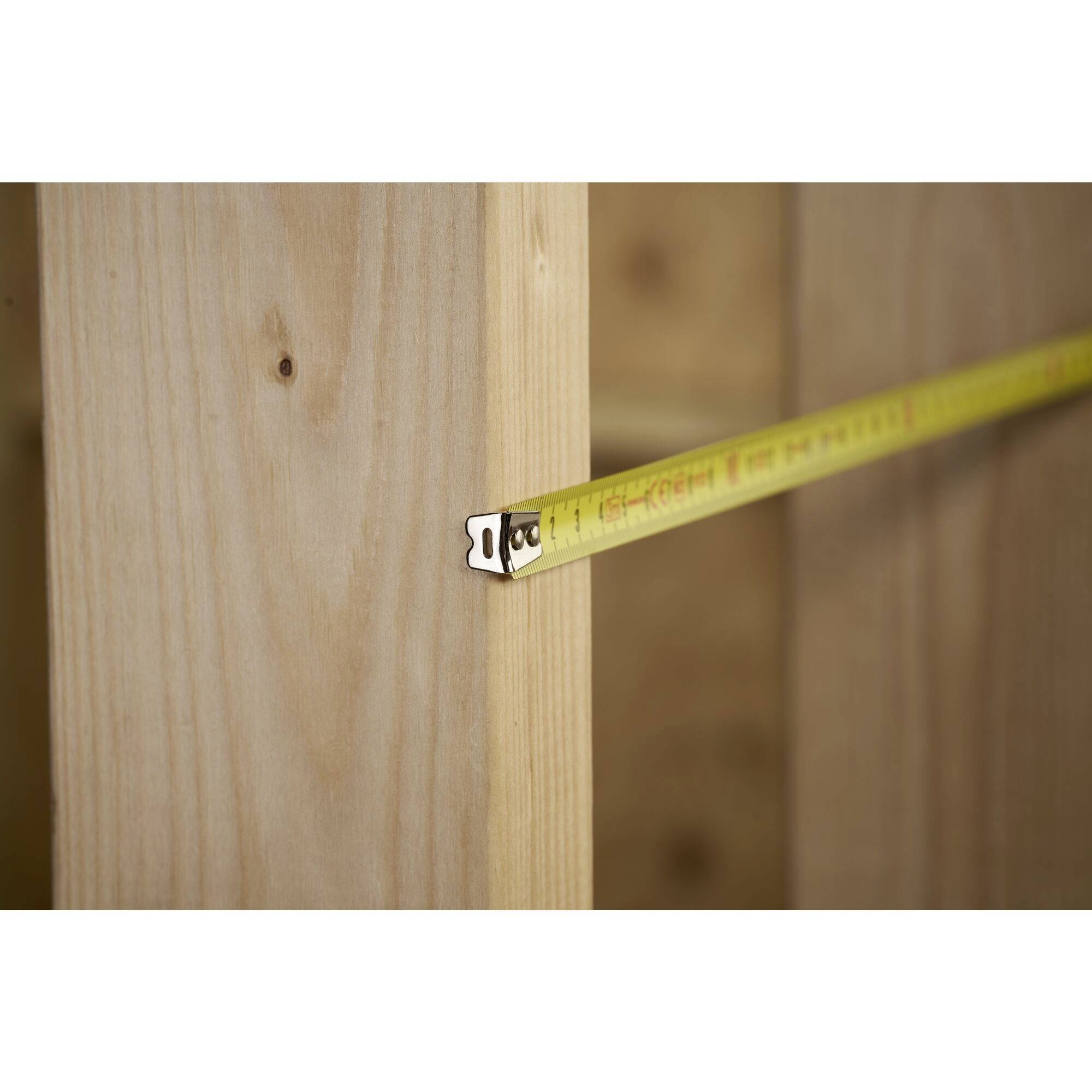 A tape measure is measuring the distance between two wooden planks. The tape measure is yellow with black markings for centimetres and inches.