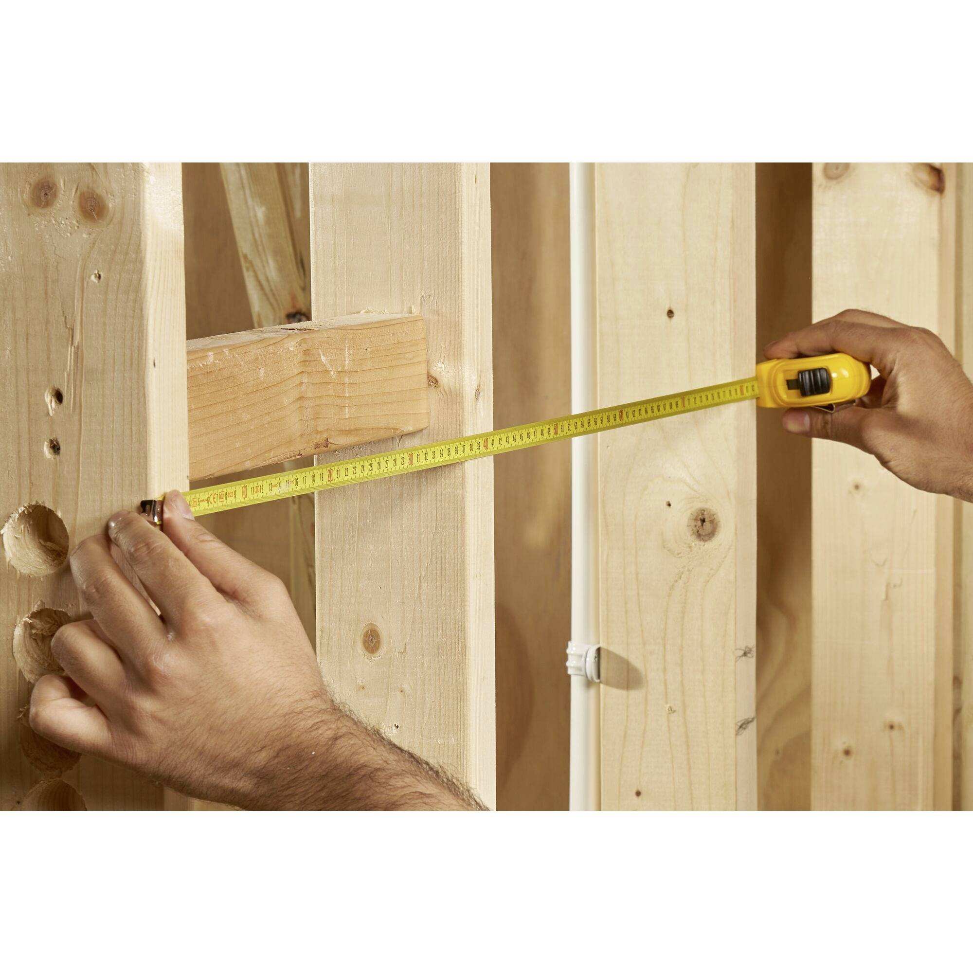 A person is measuring a wooden frame in an unfinished building using a yellow tape measure.