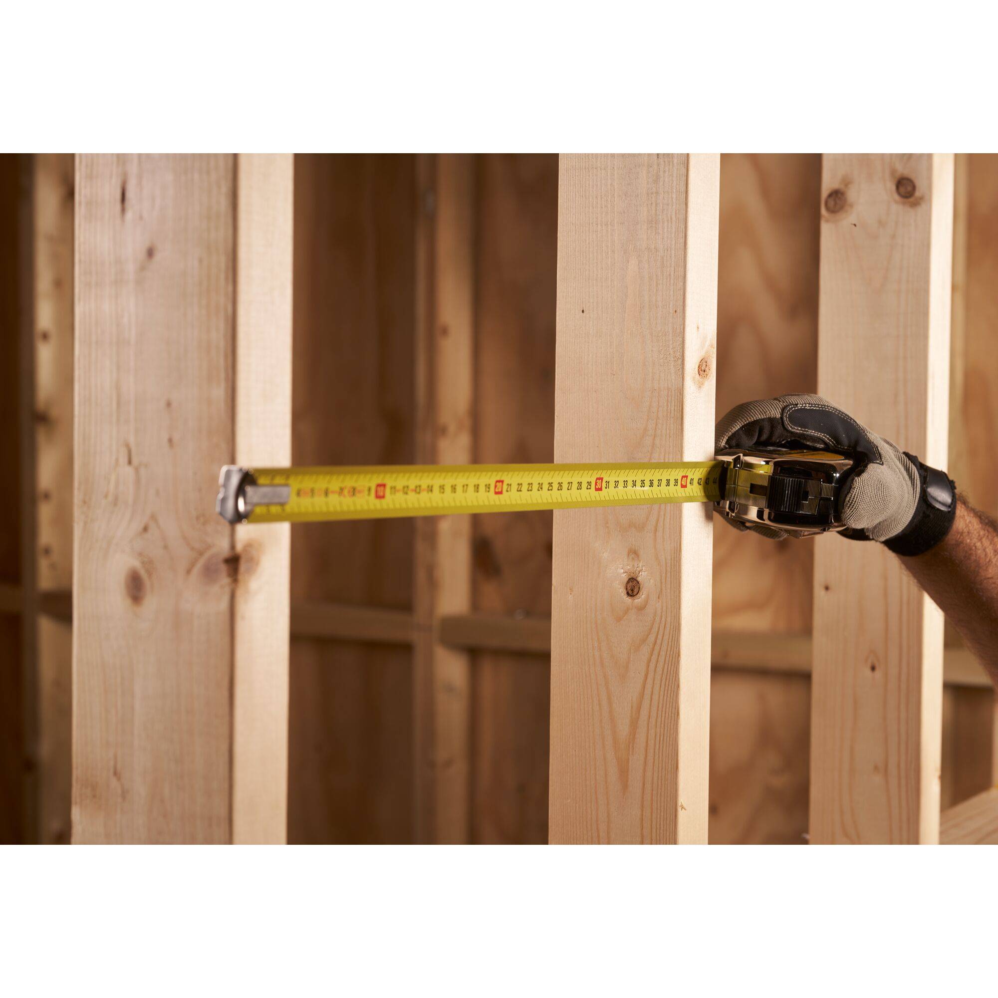 A hand wearing a glove holds a measuring tape, measuring a wooden beam in an unfinished construction wall.