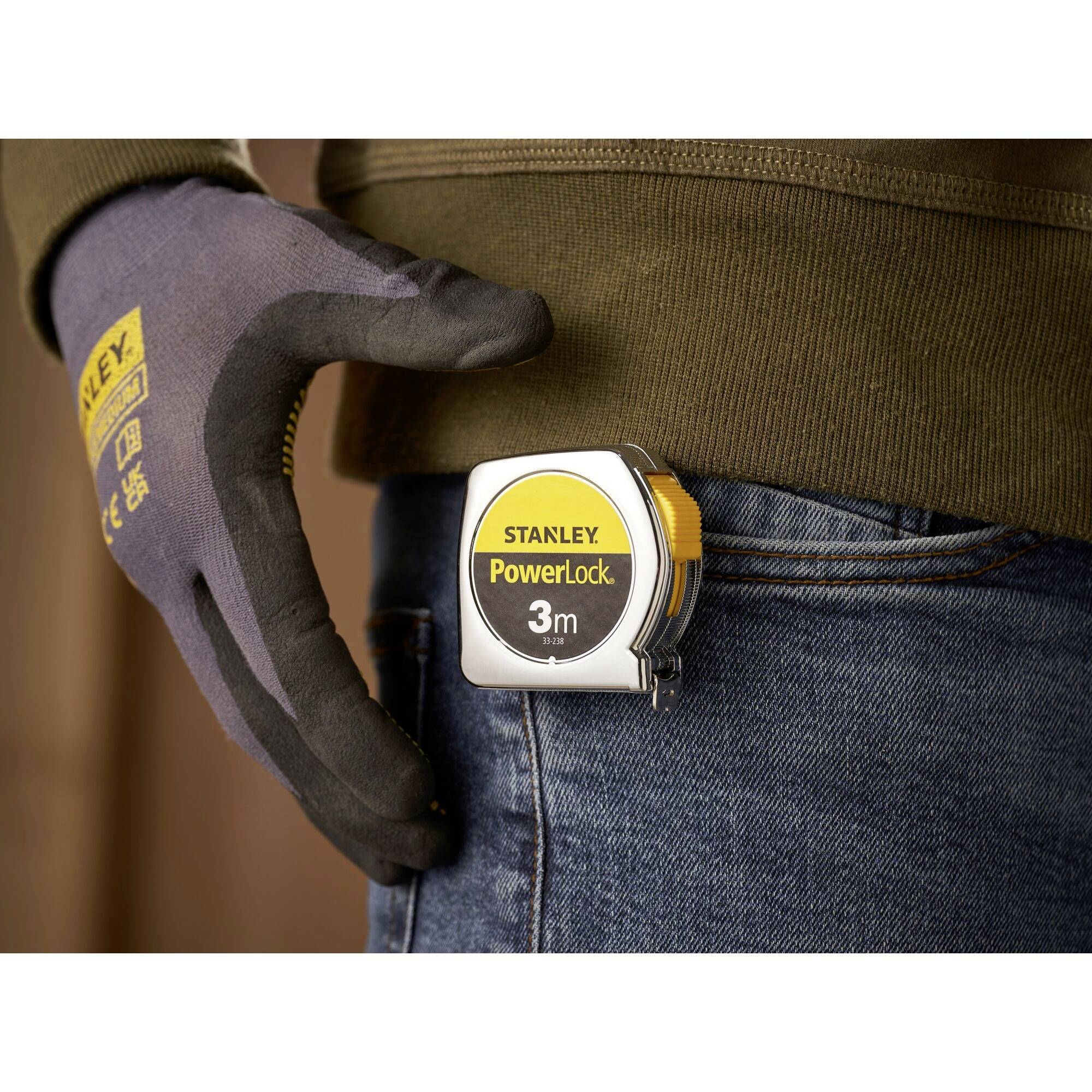 A person is wearing a Stanley PowerLock tape measure on their trouser pocket. The hand is wearing a work glove.